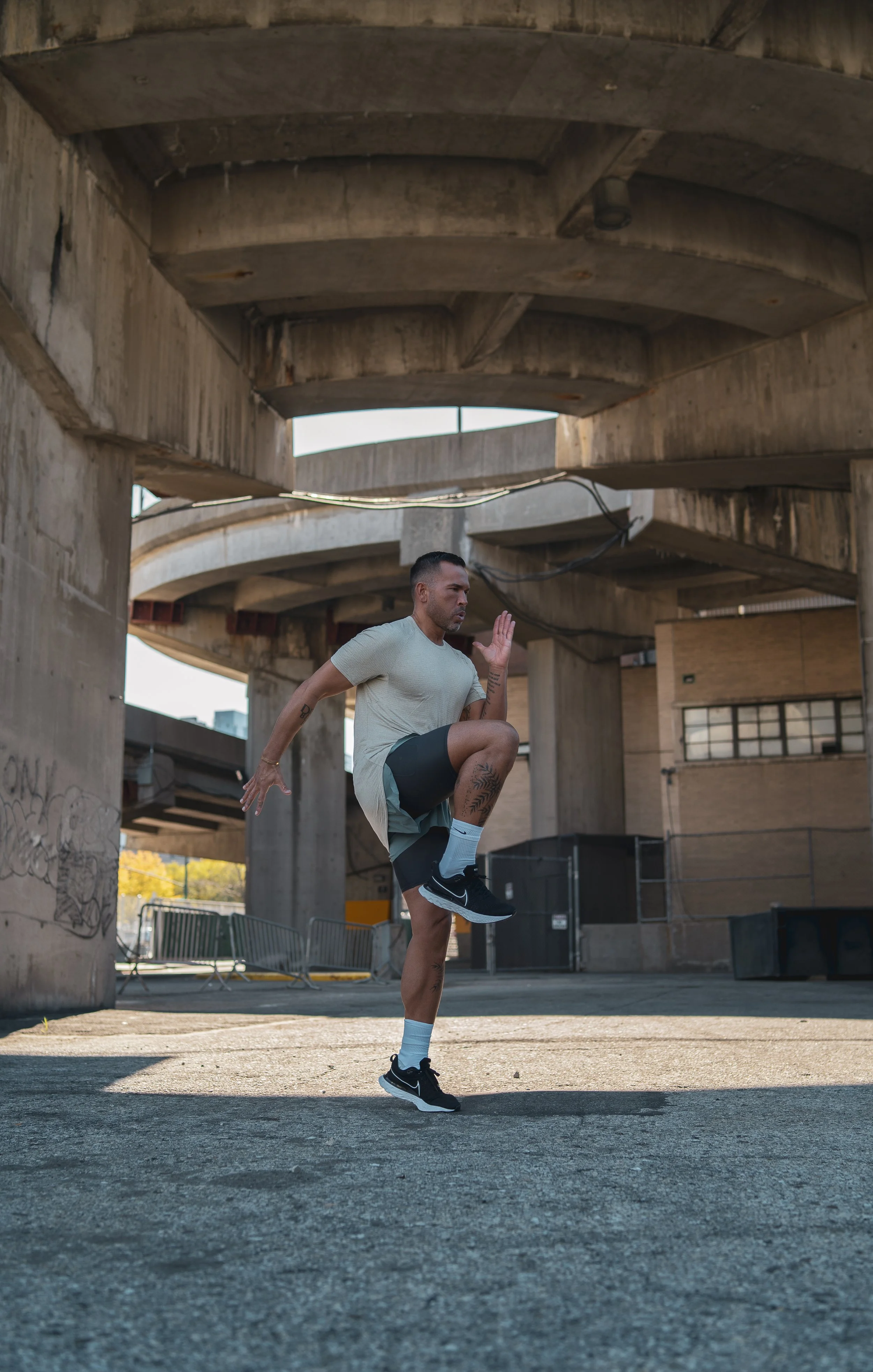 A man in athletic clothing is performing a high knee exercise under a concrete overpass.