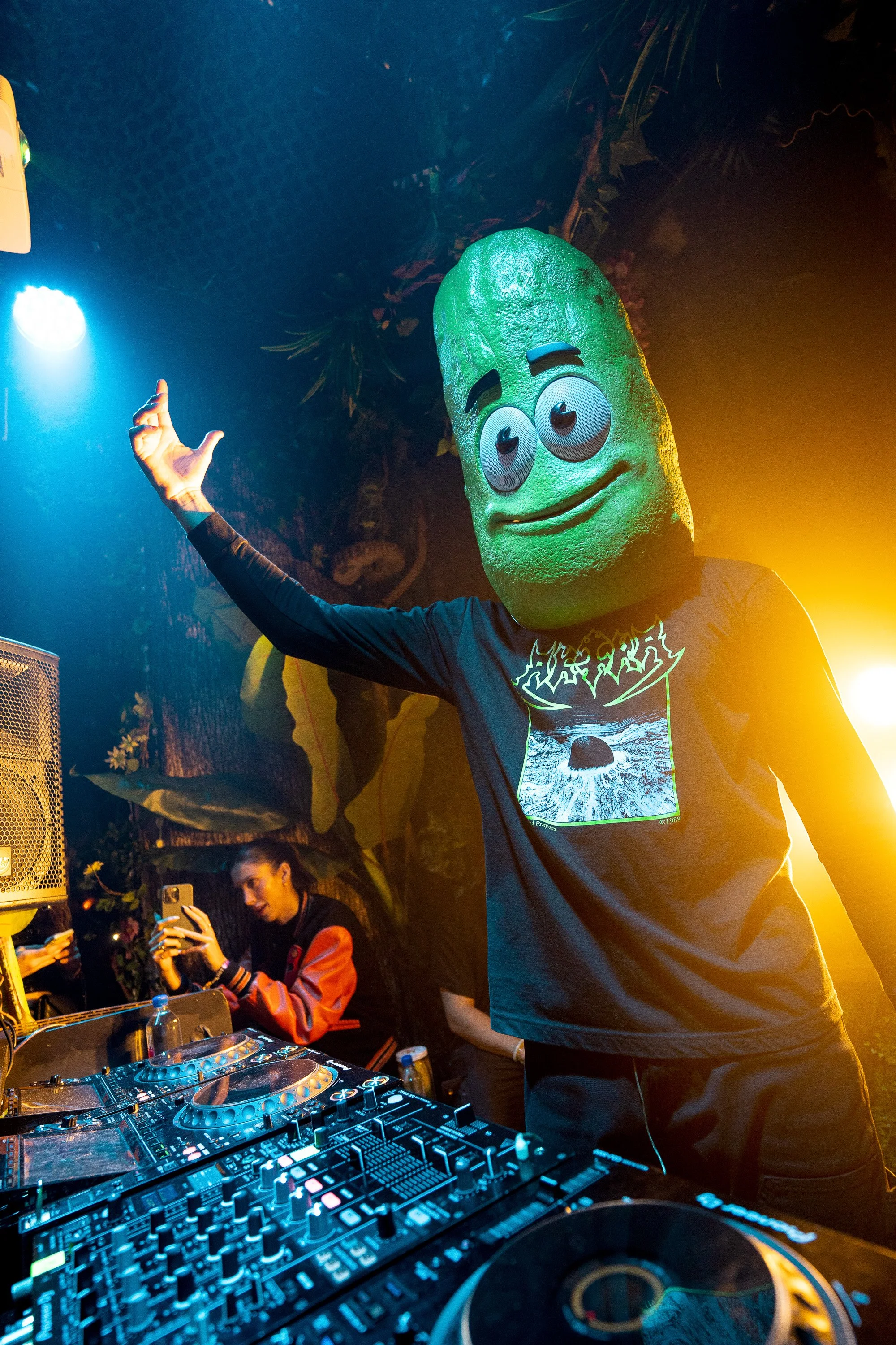 Person wearing a large green pickle mascot head DJing at a party with colorful lights.