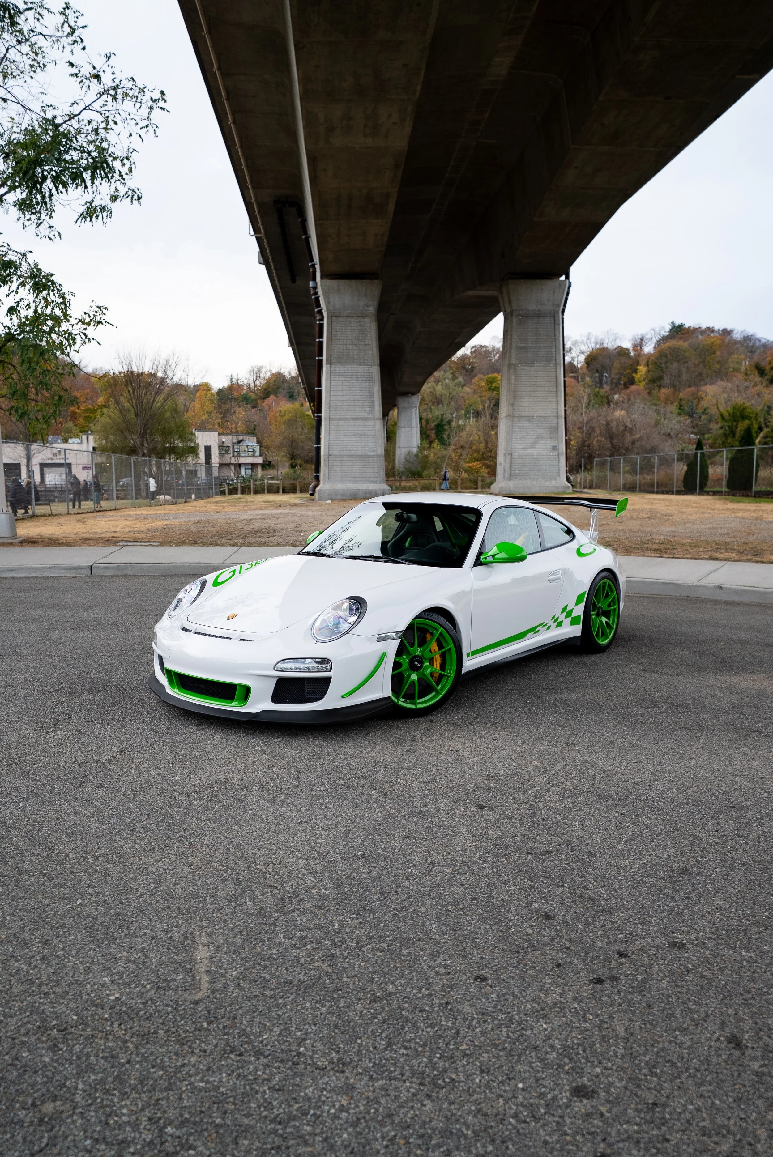 A white Porsche sports car with green accents, green wheels, and a rear spoiler parked on a street under a large bridge, with trees and a fenced area in the background during daytime.