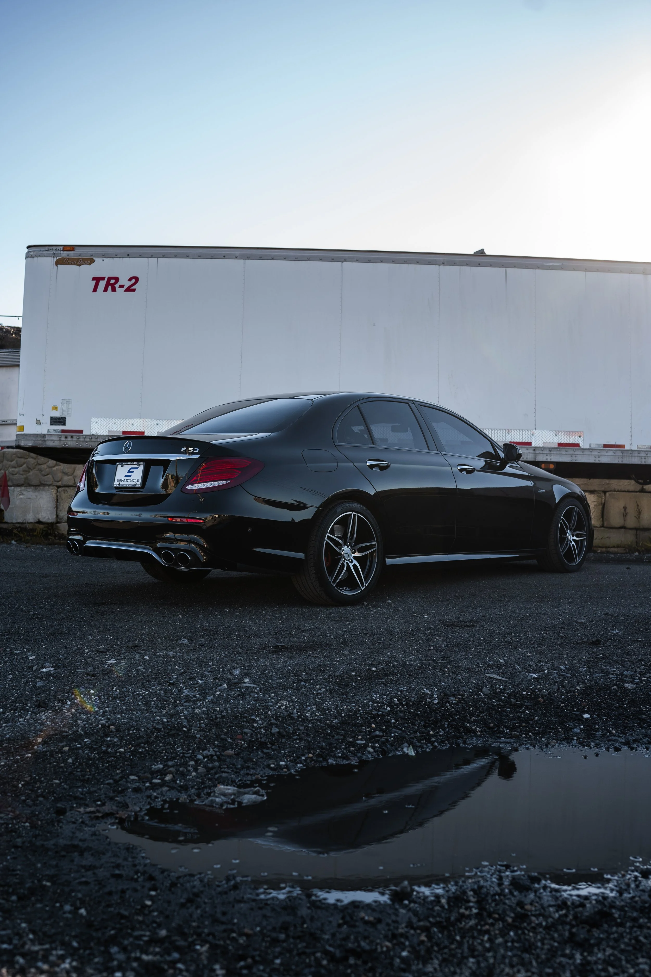 A black Mercedes-Benz E53 AMG sedan parked on a gravel surface with puddles, with a large white trailer in the background and the sky clear and bright.