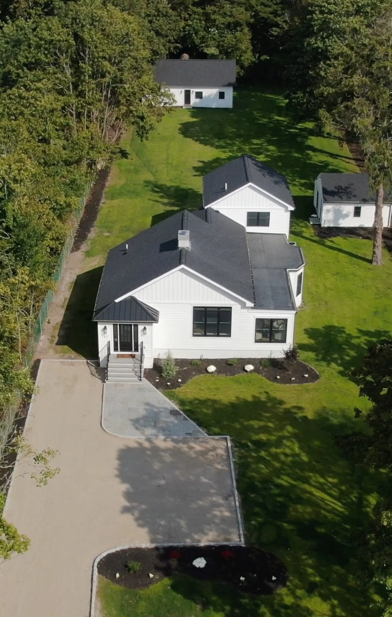 Aerial view of a modern white house with black window frames, surrounded by a well-manicured lawn, trees, and small outbuildings in a green, wooded area.