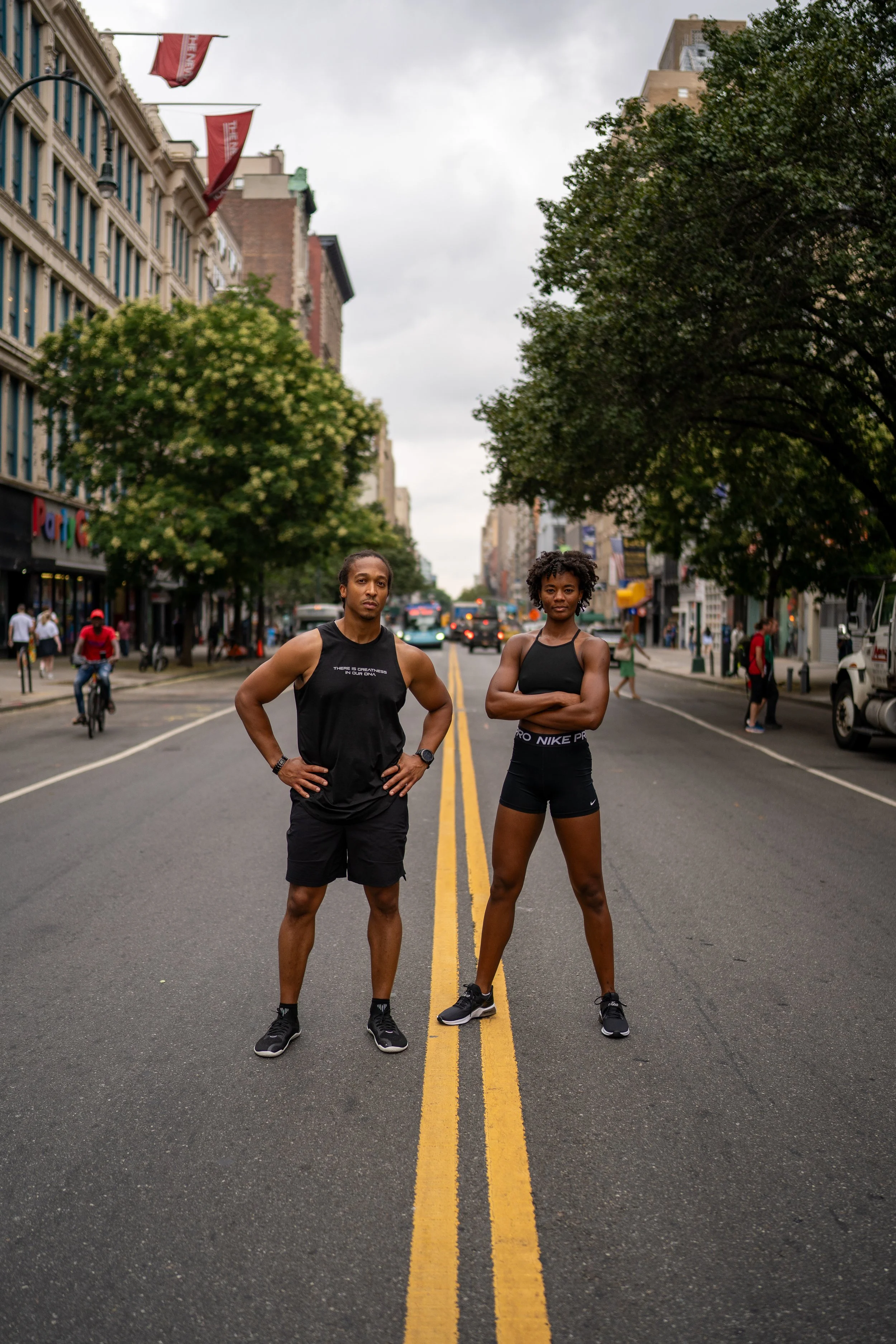 Two young athletes, a man and a woman, standing in the middle of a city street with buildings, trees, and pedestrians in the background on a cloudy day.