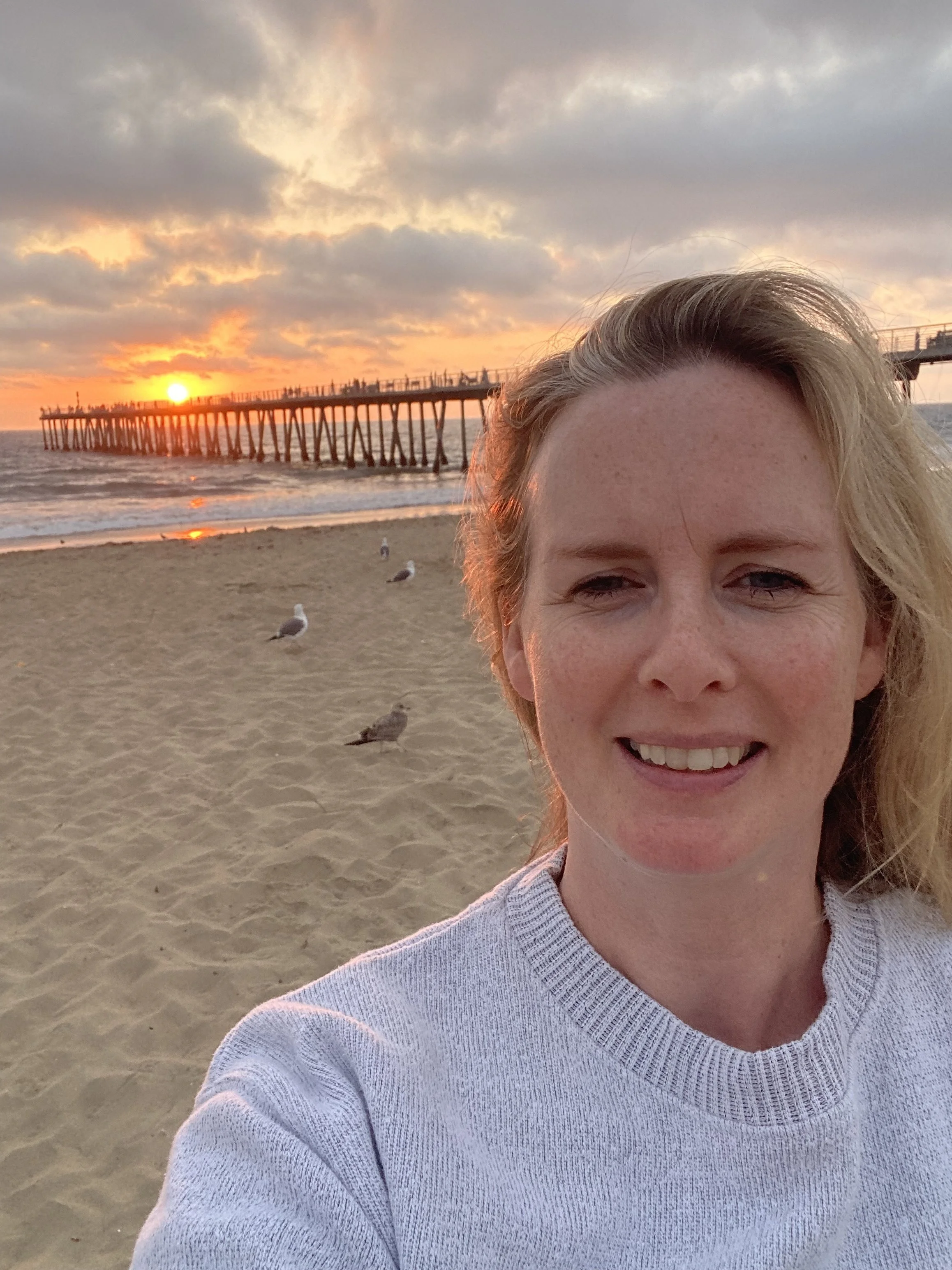 A woman taking a selfie at the beach during sunset, with seagulls on the sand and a pier extending into the ocean in the background.