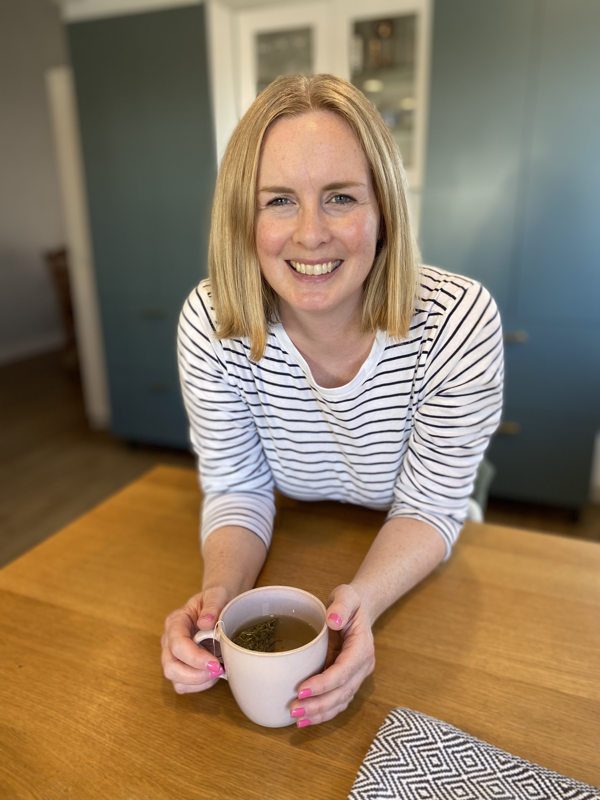 A smiling woman with blonde hair in a striped shirt is sitting at a wooden table, receiving a cup of tea or coffee from another person's hand. In the background, there are blue and white cabinets, and a gray wall. The woman has pink nail polish.