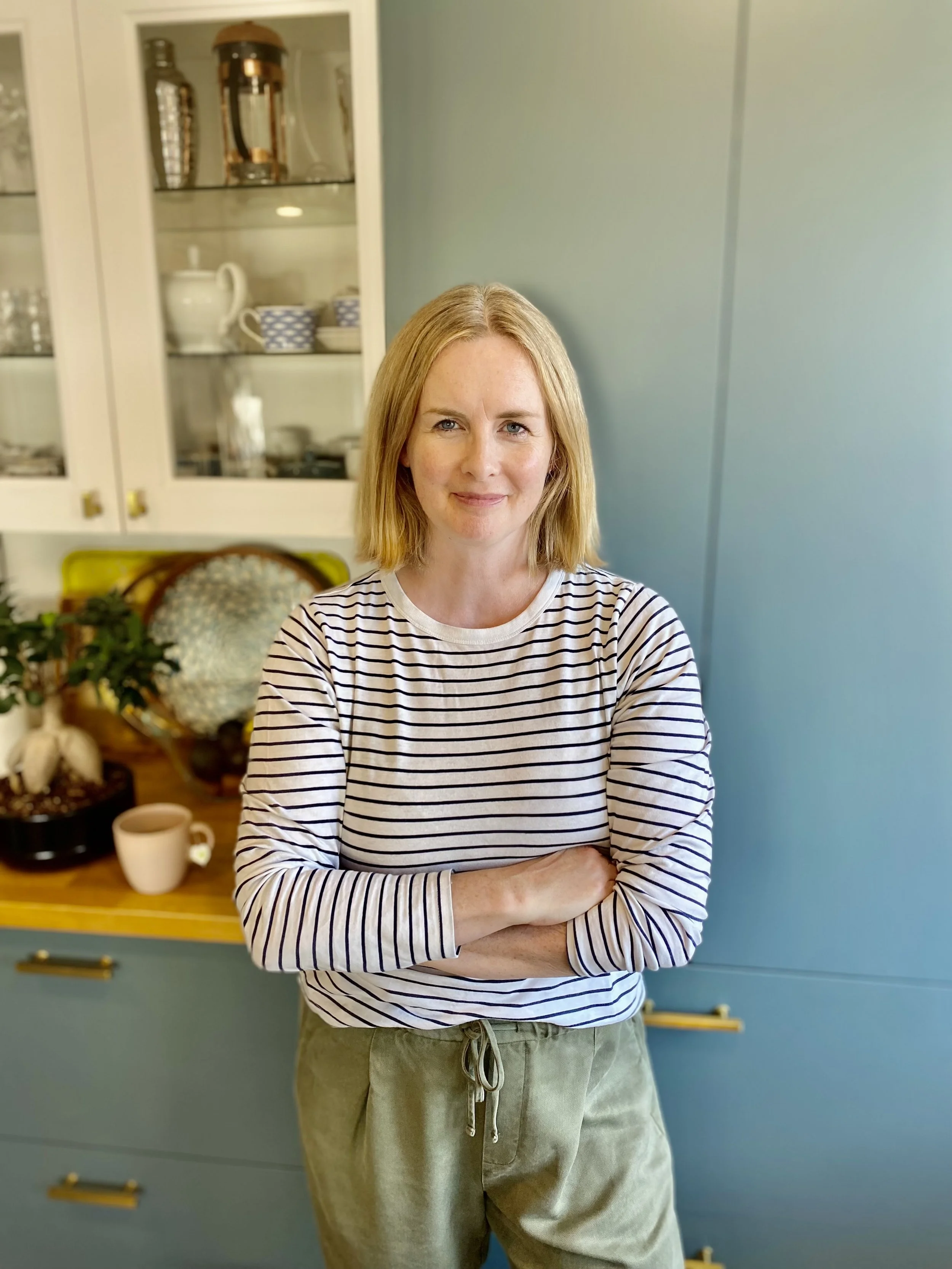 A woman with shoulder-length blonde hair, wearing a white and black striped long-sleeve shirt and light-green pants, standing with her arms crossed in a kitchen with a blue wall and a white cabinet with glass doors in the background.