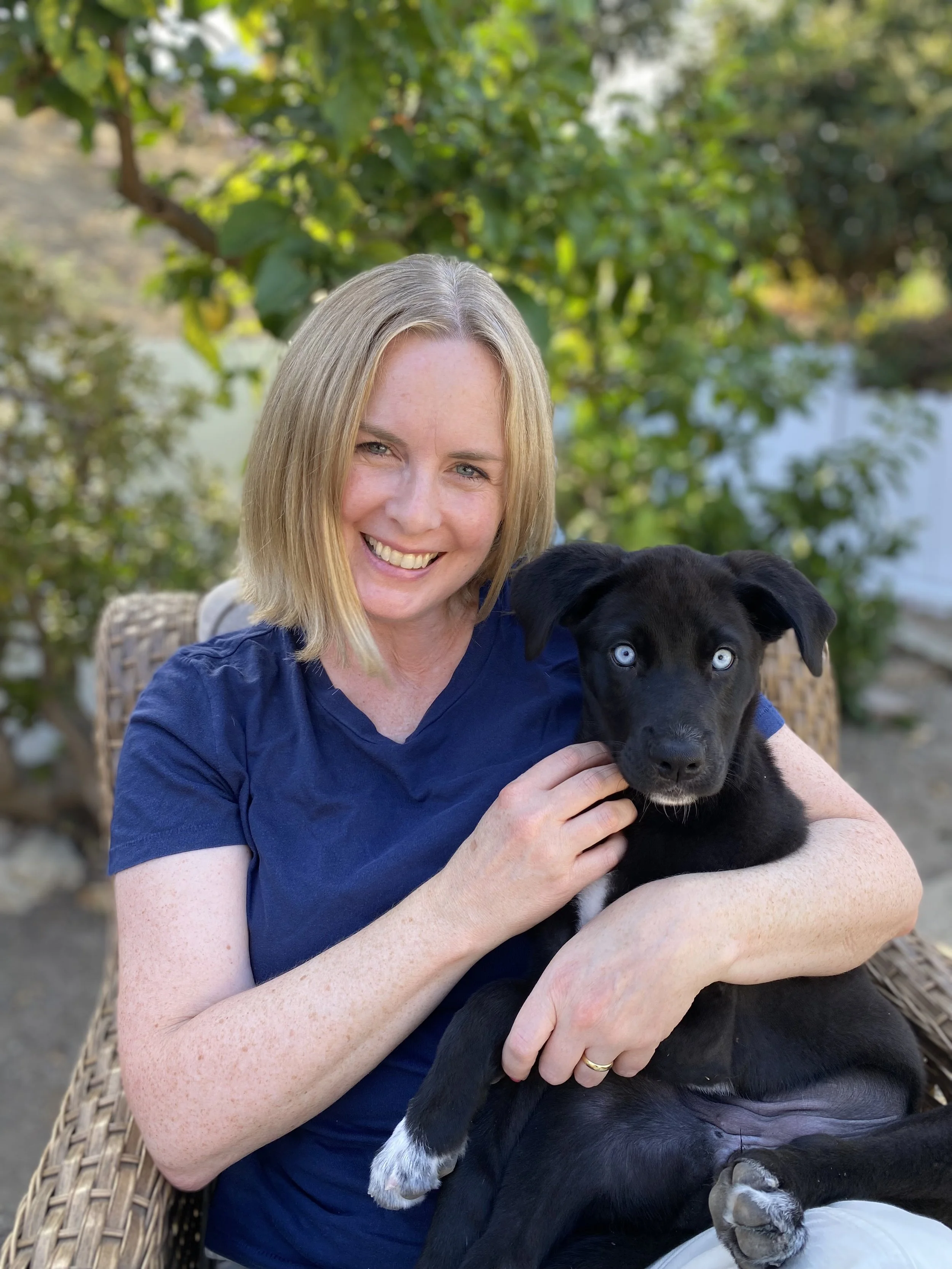 A woman with blonde hair and a blue shirt holding a black puppy with blue eyes, sitting outdoors with green foliage in the background.