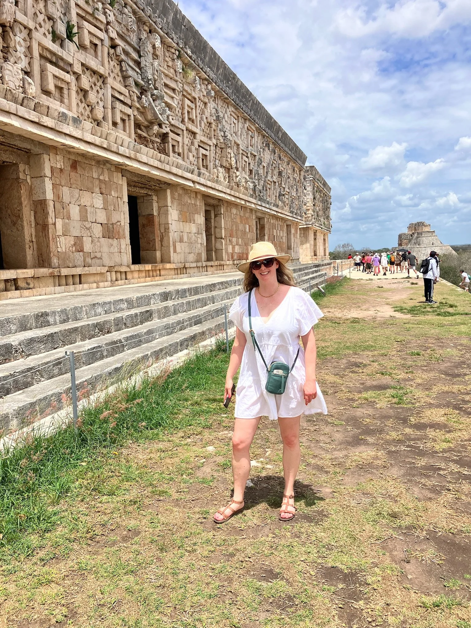 A woman standing outdoors near ancient stone ruins, wearing a white dress, sandals, a straw hat, and sunglasses, smiling at the camera with a group of tourists and ruins in the background under a partly cloudy sky.