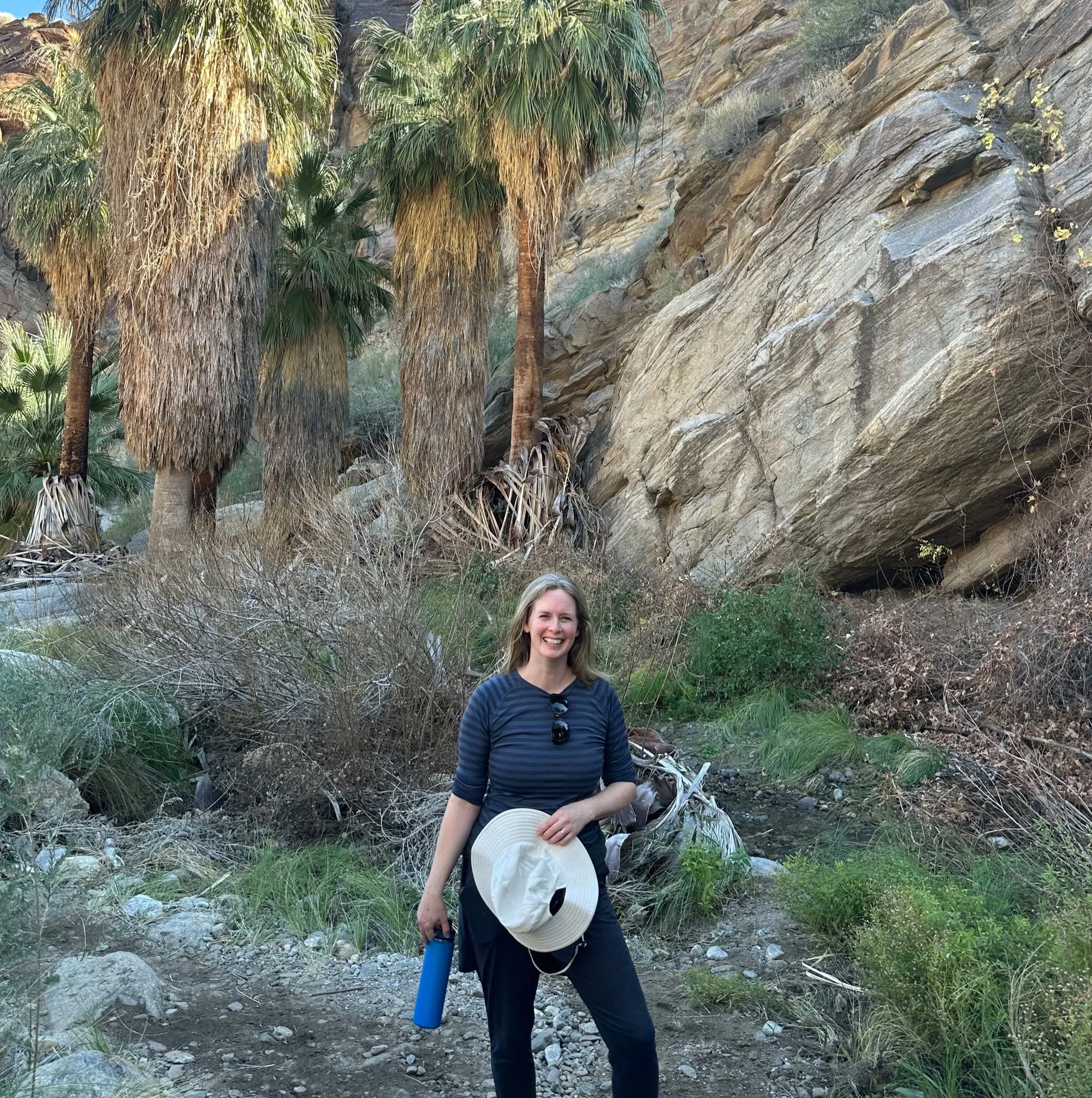 A woman standing outdoors on rocky terrain with large trees and a rocky hillside in the background. She is smiling, holding a hat in one hand, and has sunglasses hanging from her shirt collar.