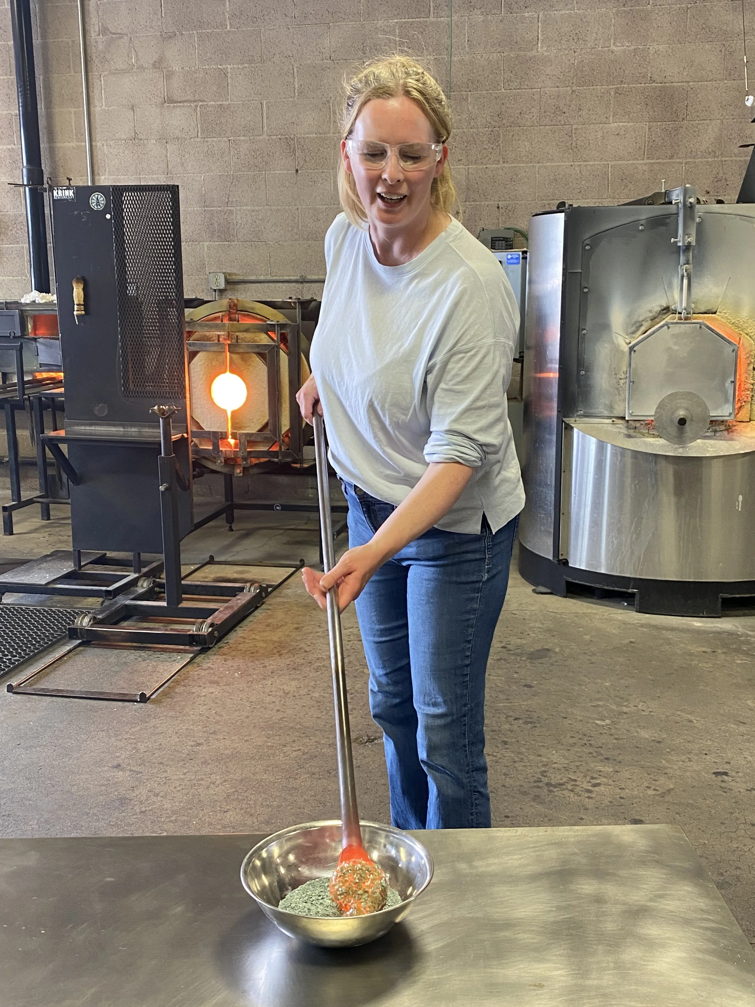 A woman wearing safety glasses and a white shirt is pouring liquid metal from a crucible into a bowl in a foundry or metalworking shop, with industrial equipment in the background.