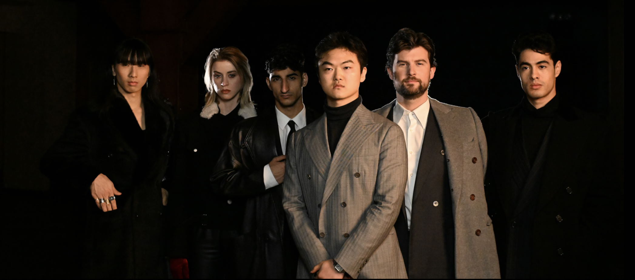 Six diverse young adults standing together indoors, dressed in stylish clothing, with serious and confident expressions, against a dark background.