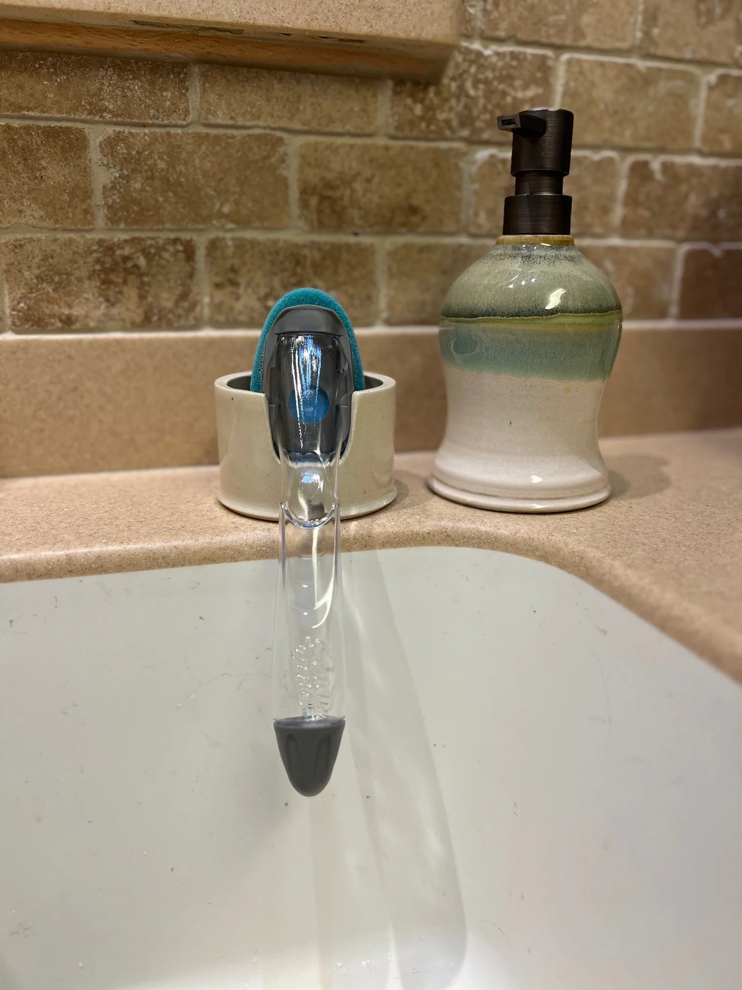 A water faucet with a blue and gray sponge resting on its base, a soap dispenser with a black pump, and a beige countertop with a white sink and a brick wall in the background.