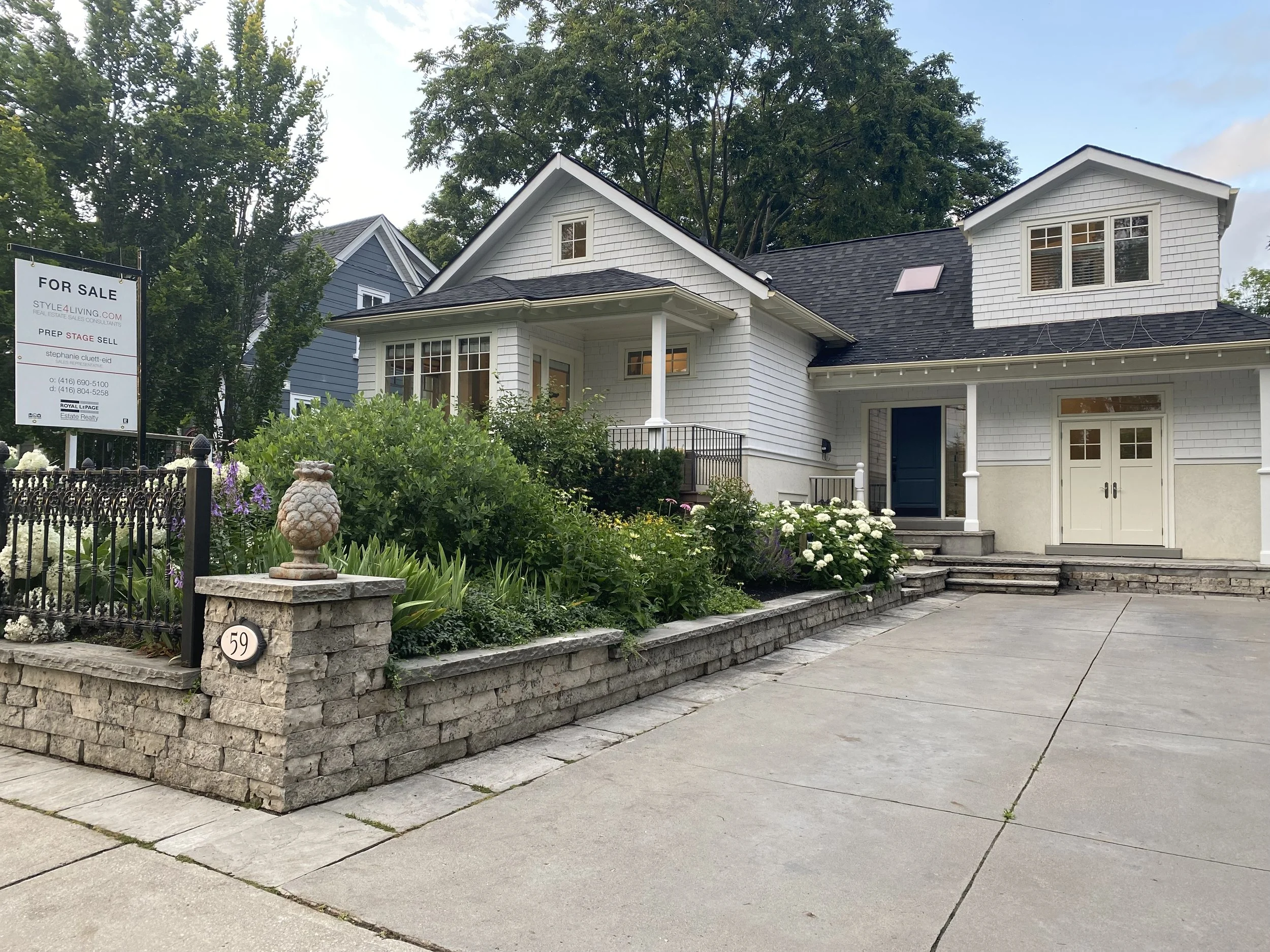 White two-story house with a dark roof, front garden with various plants, flower bed with white flowers, stone steps, driveway, and a For Sale sign in the yard