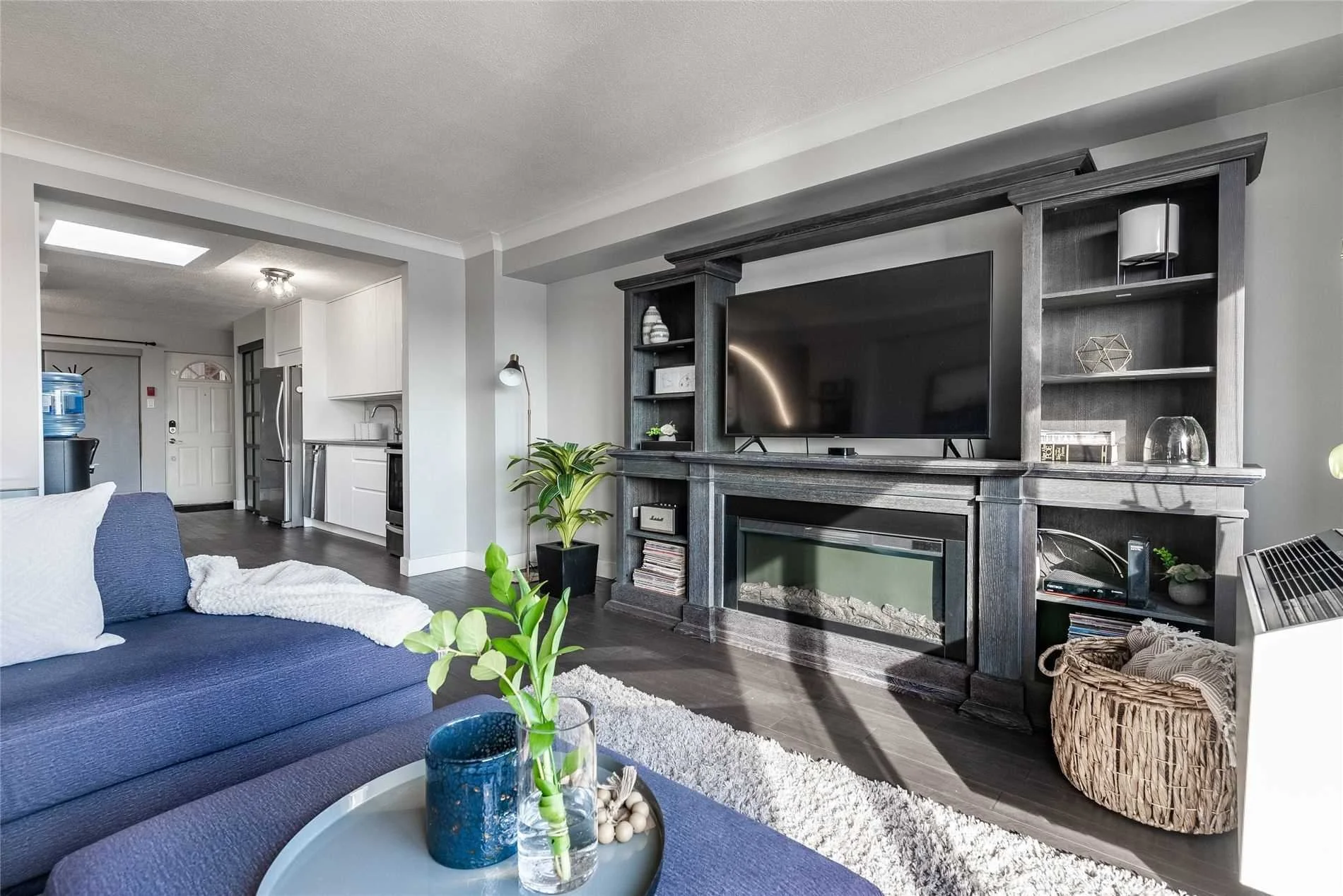 Living room with navy sofa, coffee table with green plant and decor, white area rug, and dark wood fireplace with TV above, adjacent to a kitchen with white cabinets and stainless steel appliances.