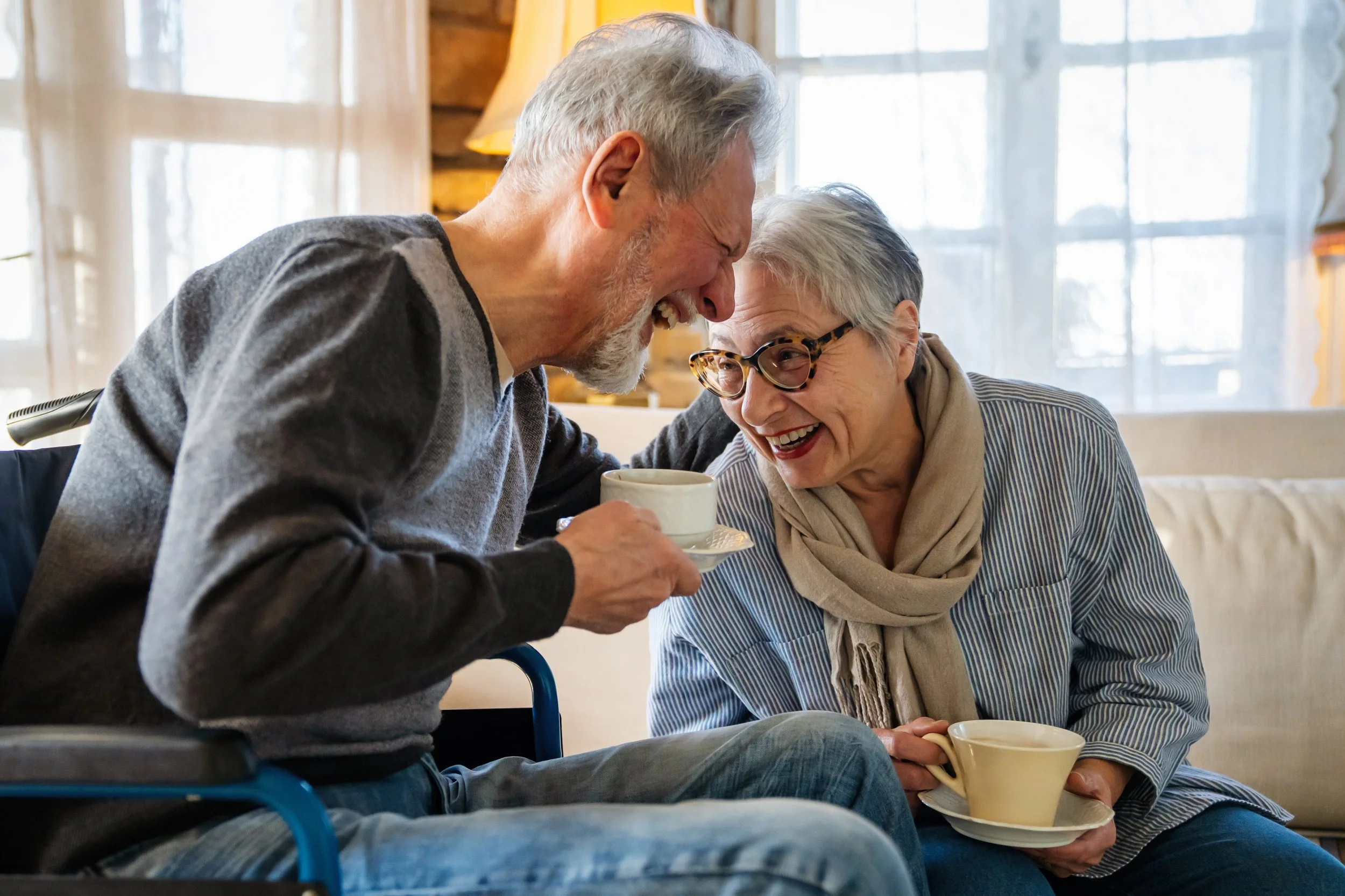 An elderly man in a wheelchair laughing and holding a cup, sharing a joyful moment with an elderly woman sitting nearby, who is also holding a cup, in a cozy room with natural light.