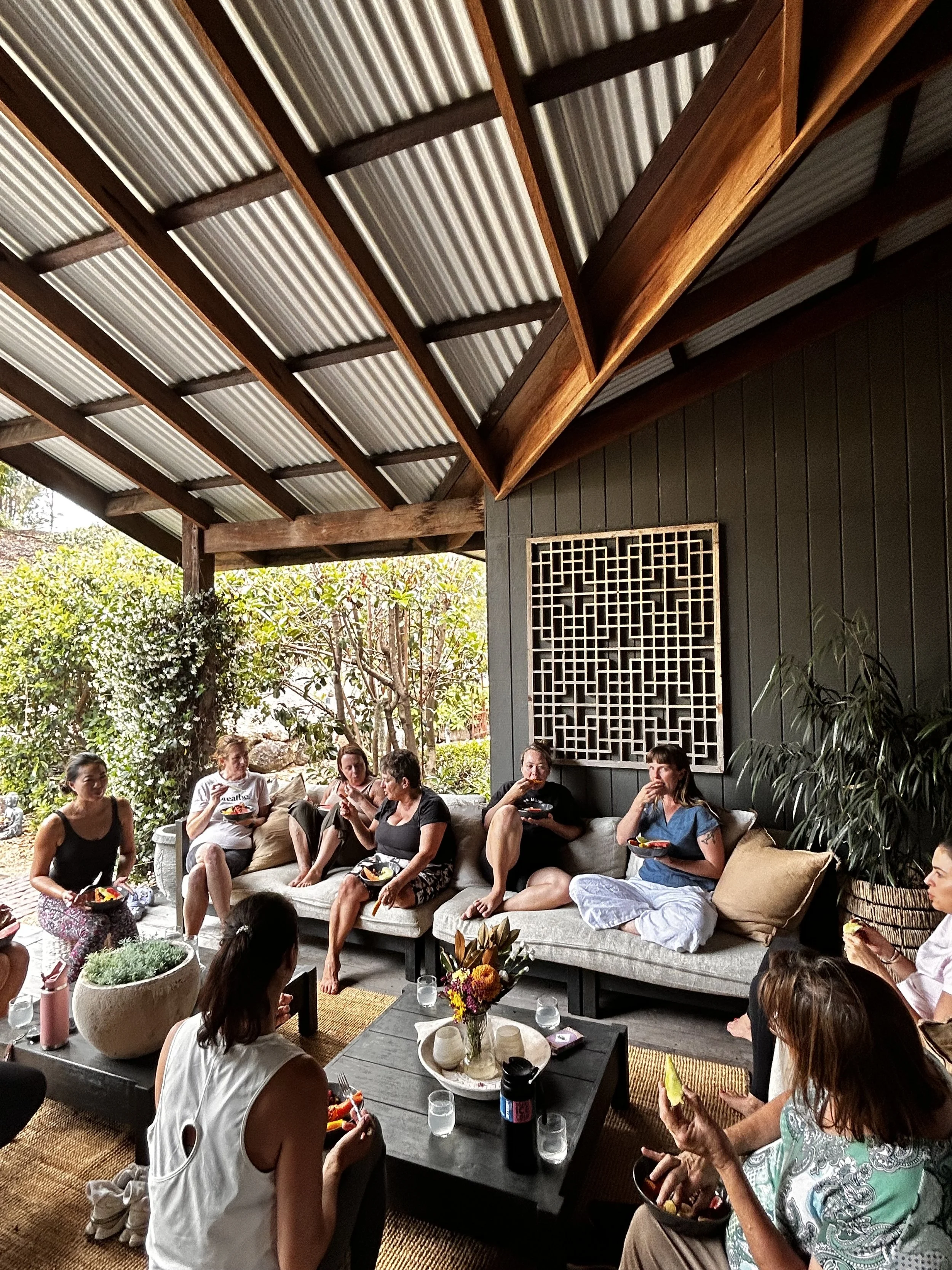 Group of women gathered on a covered outdoor patio, sitting on couches and chairs, enjoying food and conversation with a garden view in the background.