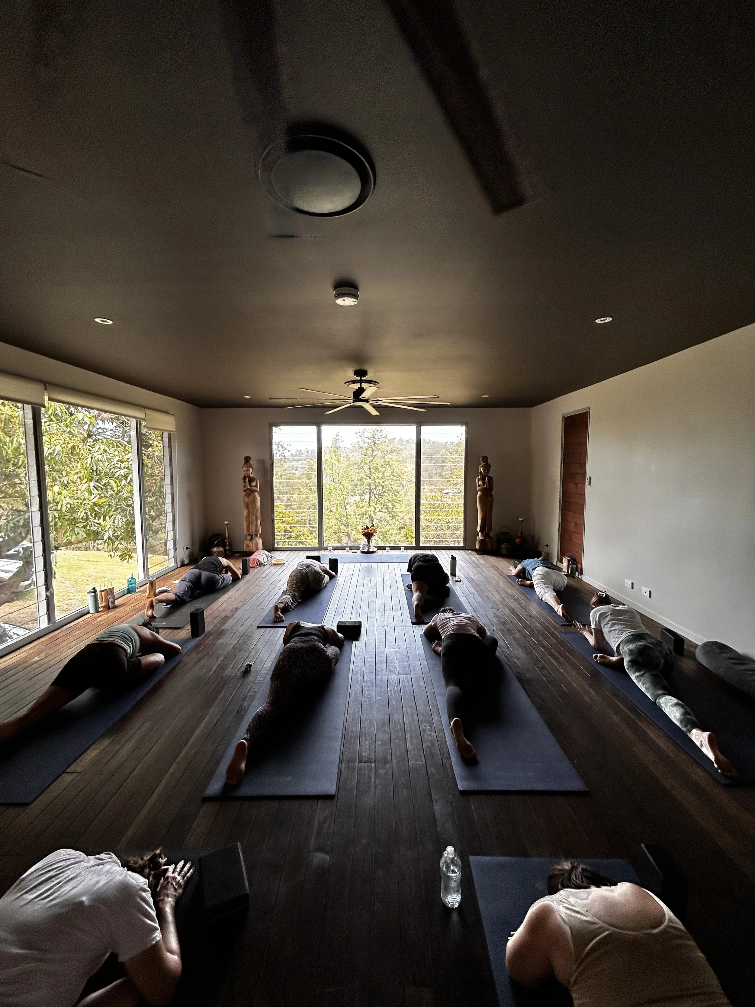 People participating in a yoga class in a spacious room with large windows and wooden floors.
