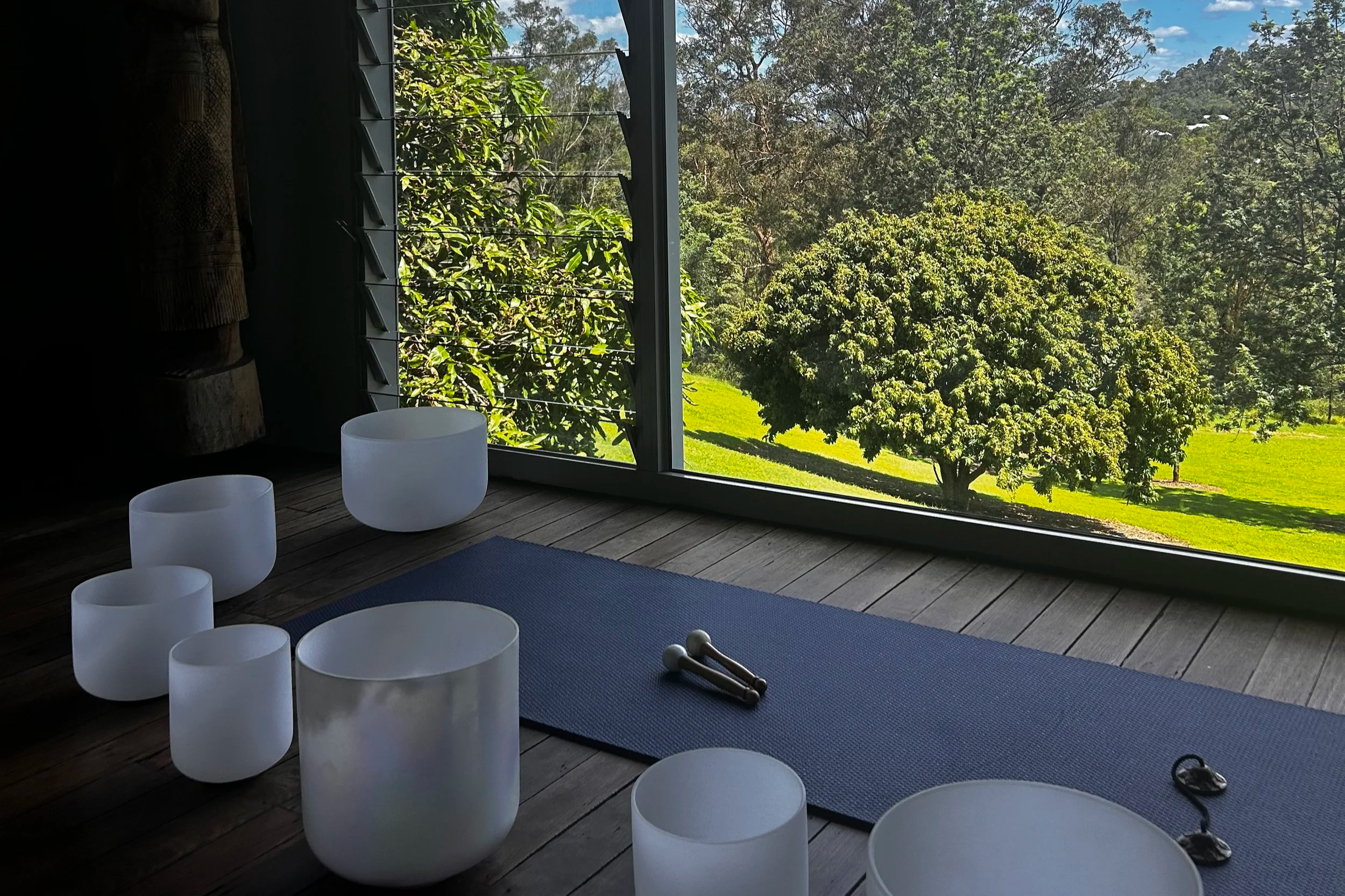 Interior of a yoga or meditation room with white crystal singing bowls on a wooden floor, a blue yoga mat, a pair of small hand weights, and a pair of glasses, with a large window overlooking a lush green landscape with trees and grass.