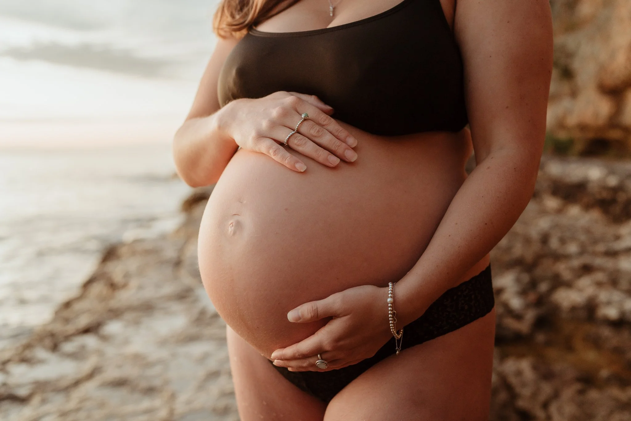 Pregnant woman in black lingerie holding her belly by the beach.