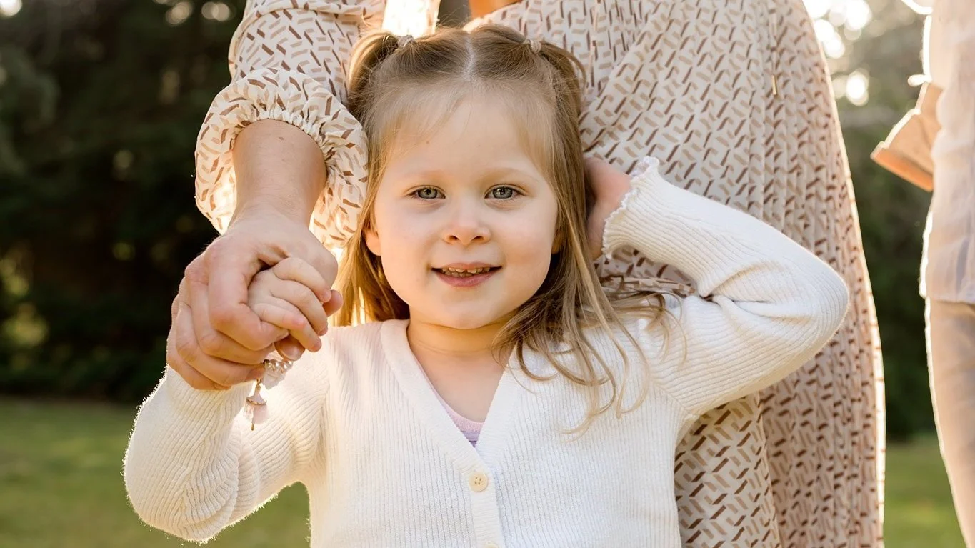A smiling young girl in a white sweater holding an adult's hand outdoors.
