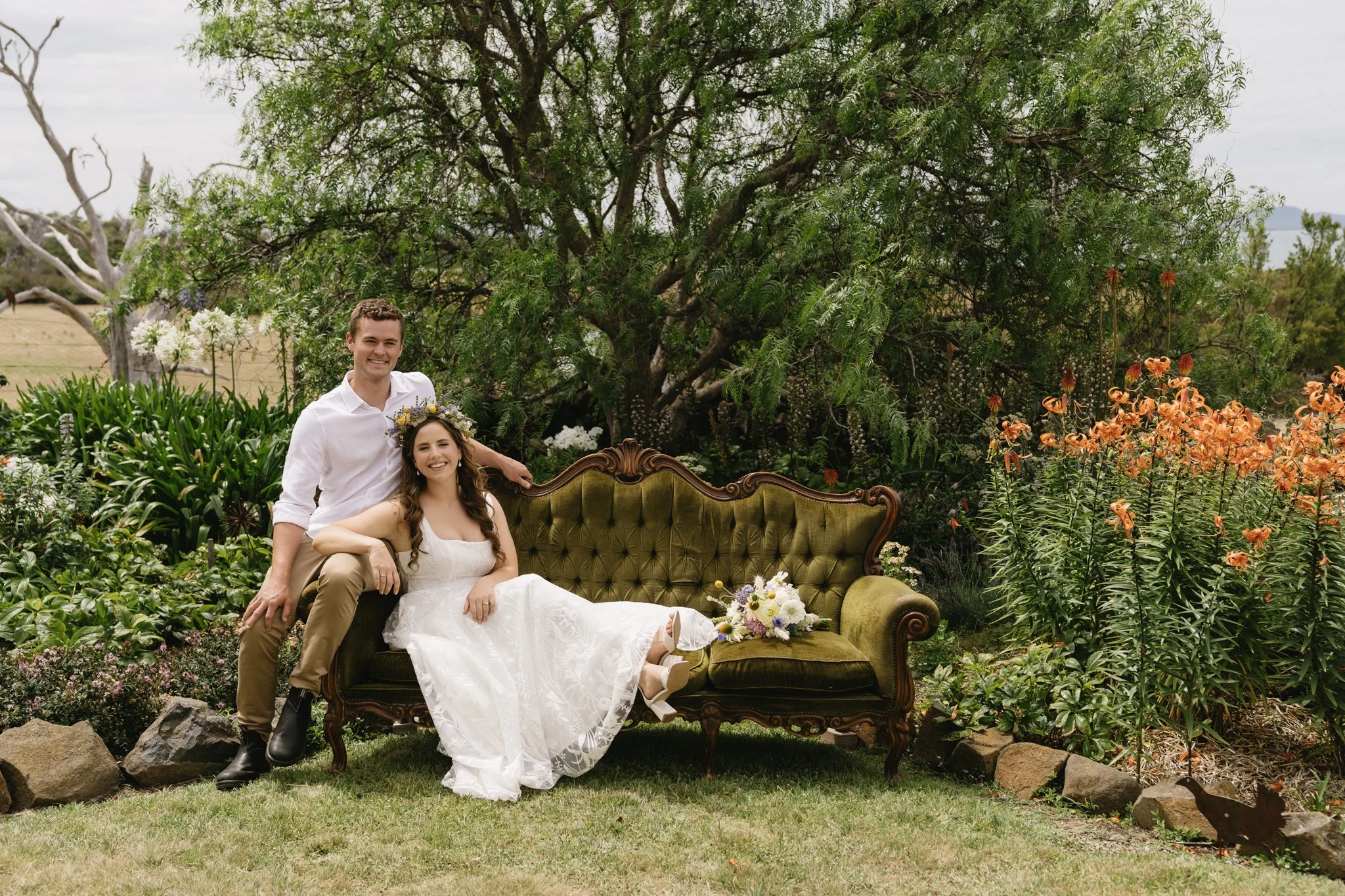Couple in garden setting, seated on a vintage green couch, surrounded by lush greenery and orange flowers.