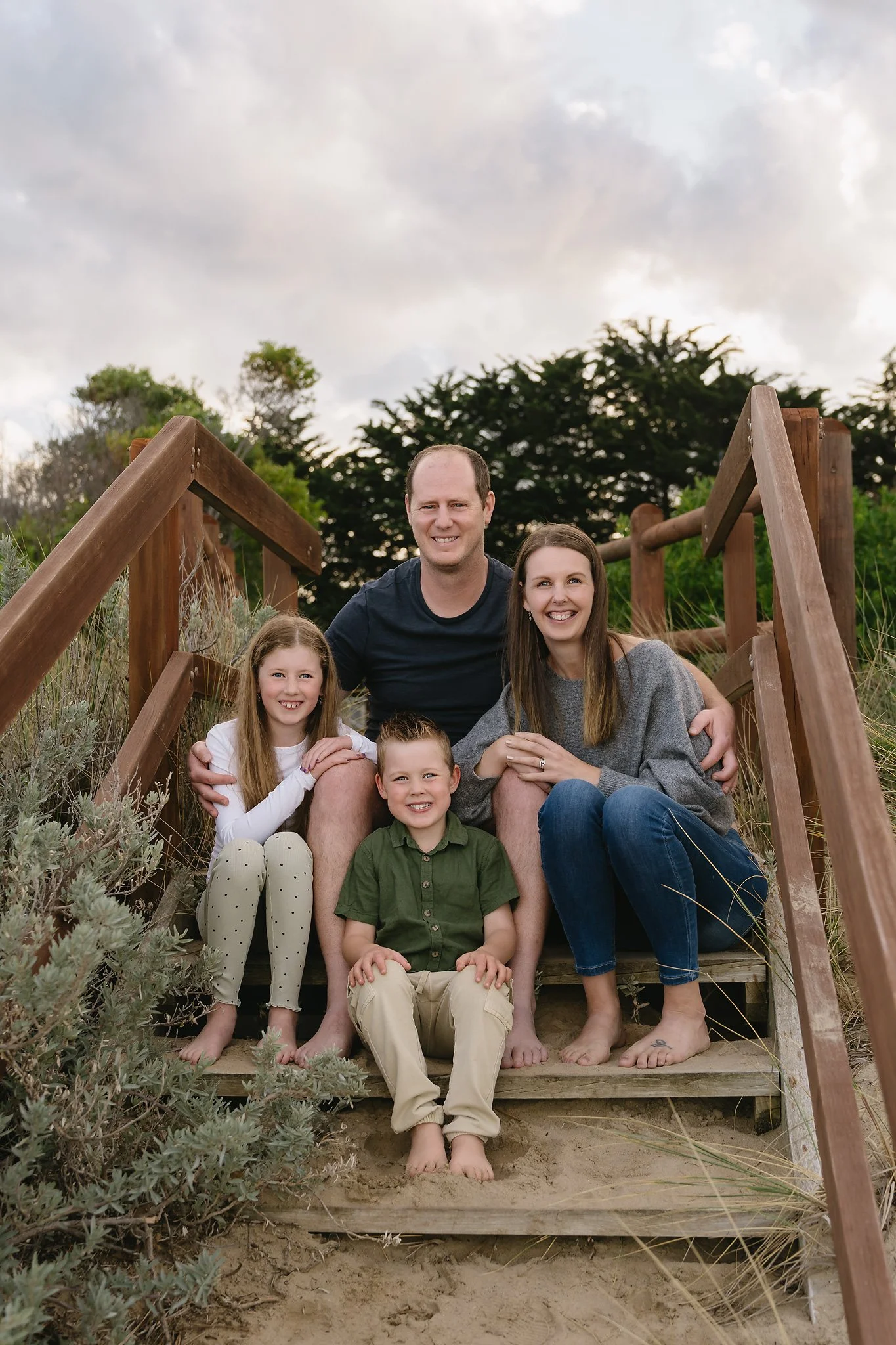 A family of five sitting barefoot on wooden stairs at a beach, smiling at the camera, with natural greenery and cloudy sky in the background.