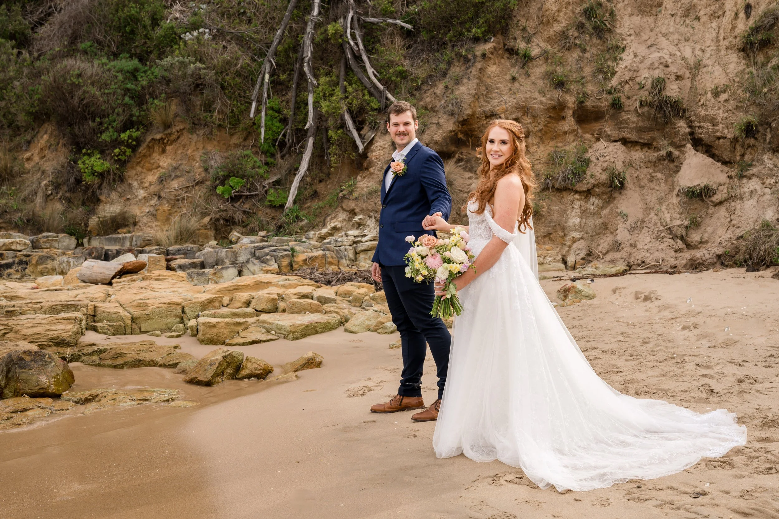 Bride and groom holding hands on a rocky beach, with bride in a white gown holding a bouquet, and groom in a dark suit.
