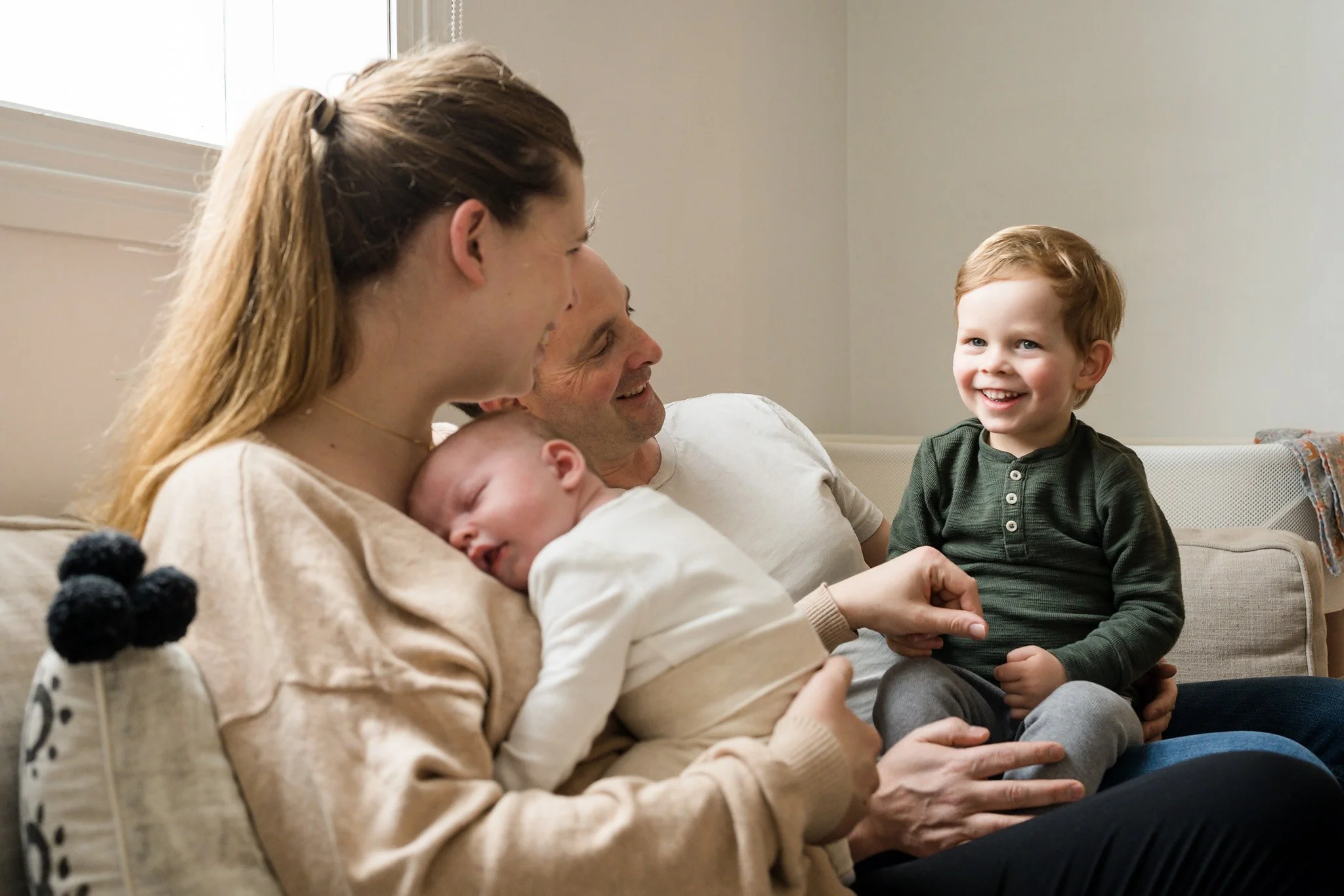 Family sitting together on a couch with a mother holding a sleeping baby and a father holding a smiling toddler.