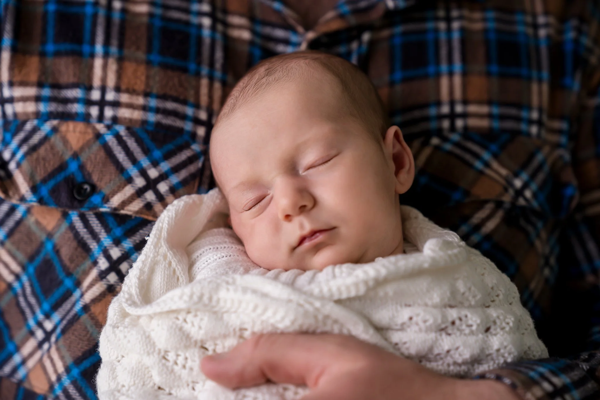 Sleeping baby wrapped in a white blanket, cradled by a person in a plaid shirt.