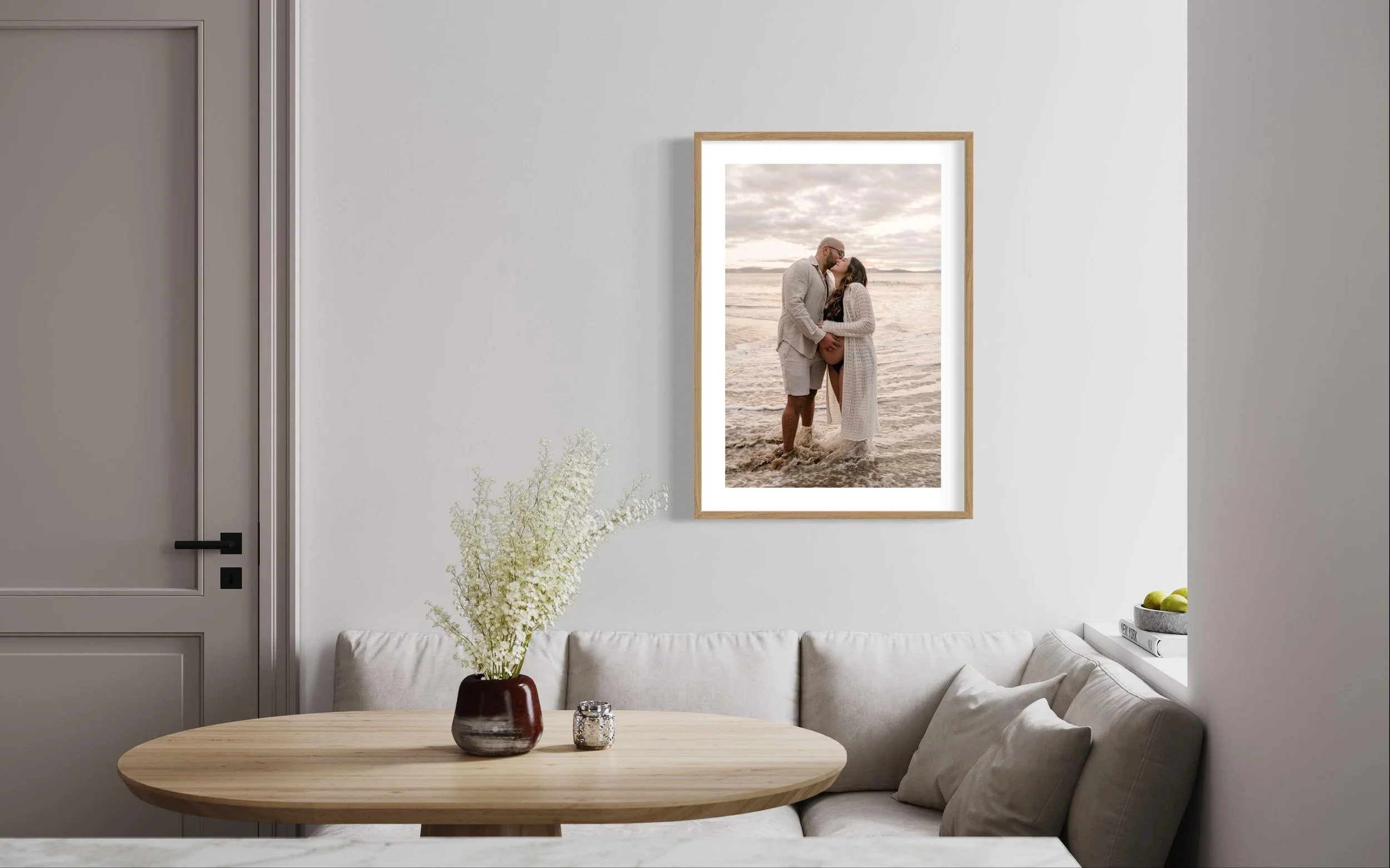 Modern living room with a wooden round table, cream-colored sofa, and a large framed photo of a couple on the beach. A vase with white flowers is on the table.