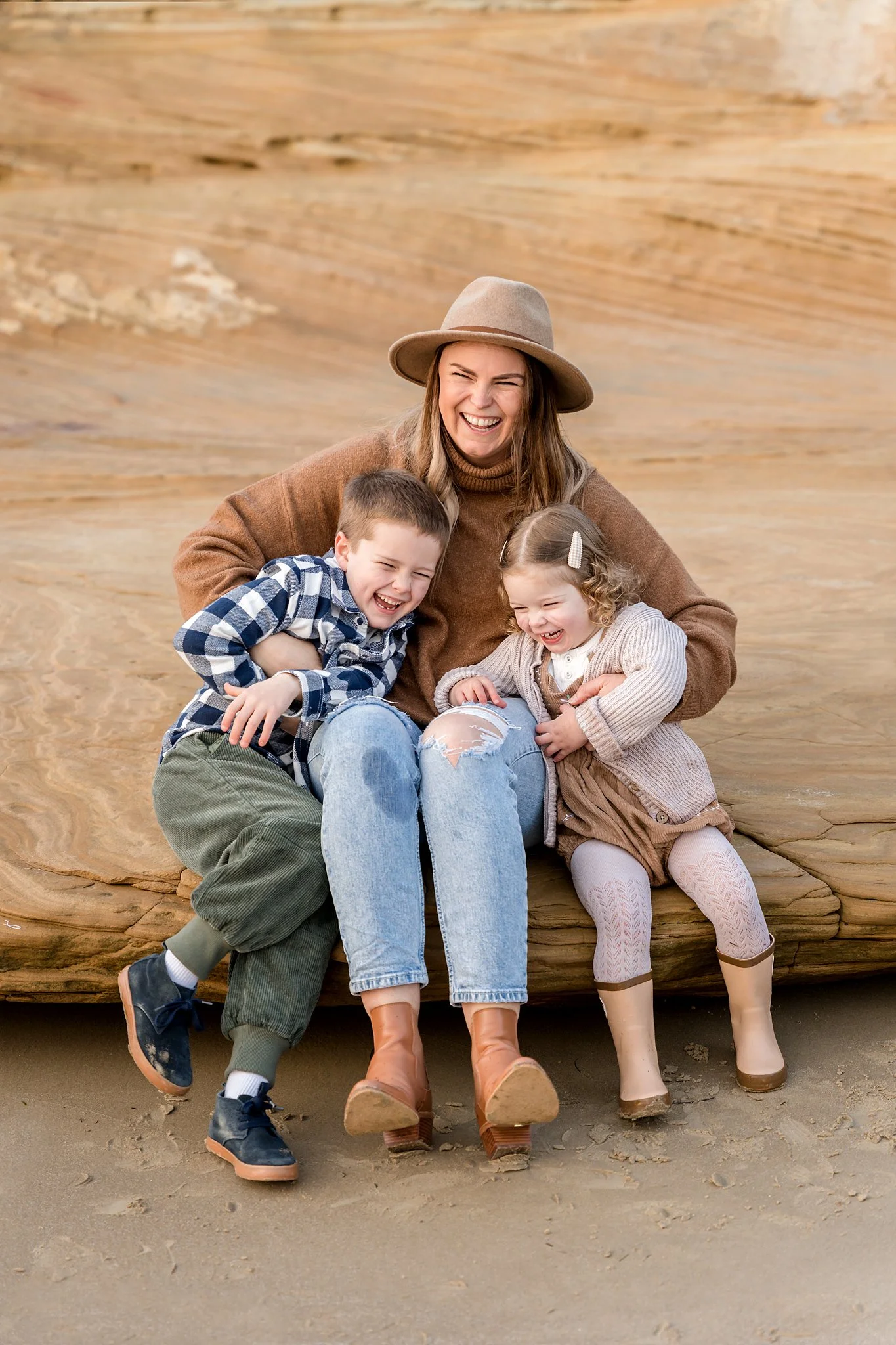 A woman in a brown sweater and hat sits on a rock with two children, a boy and a girl, both laughing. The boy is wearing a plaid shirt and pants, and the girl is wearing a cardigan and boots. The background features a sandy rock formation.