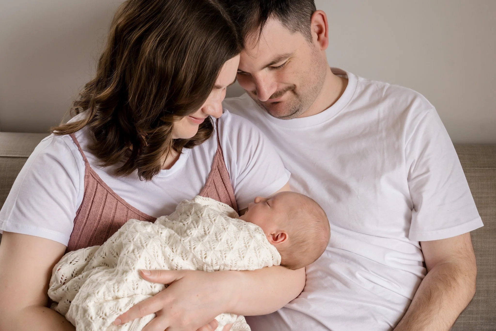 A couple in white shirts lovingly looking at their newborn baby wrapped in a knit blanket.