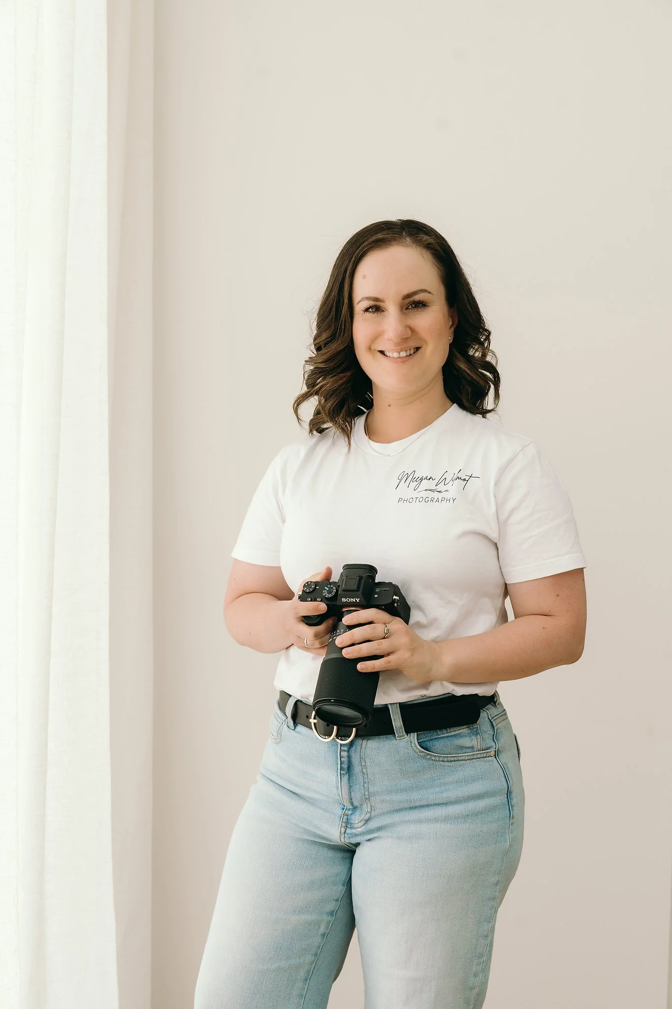 Person holding a camera, wearing a white T-shirt and jeans, smiling, standing in front of a light background.