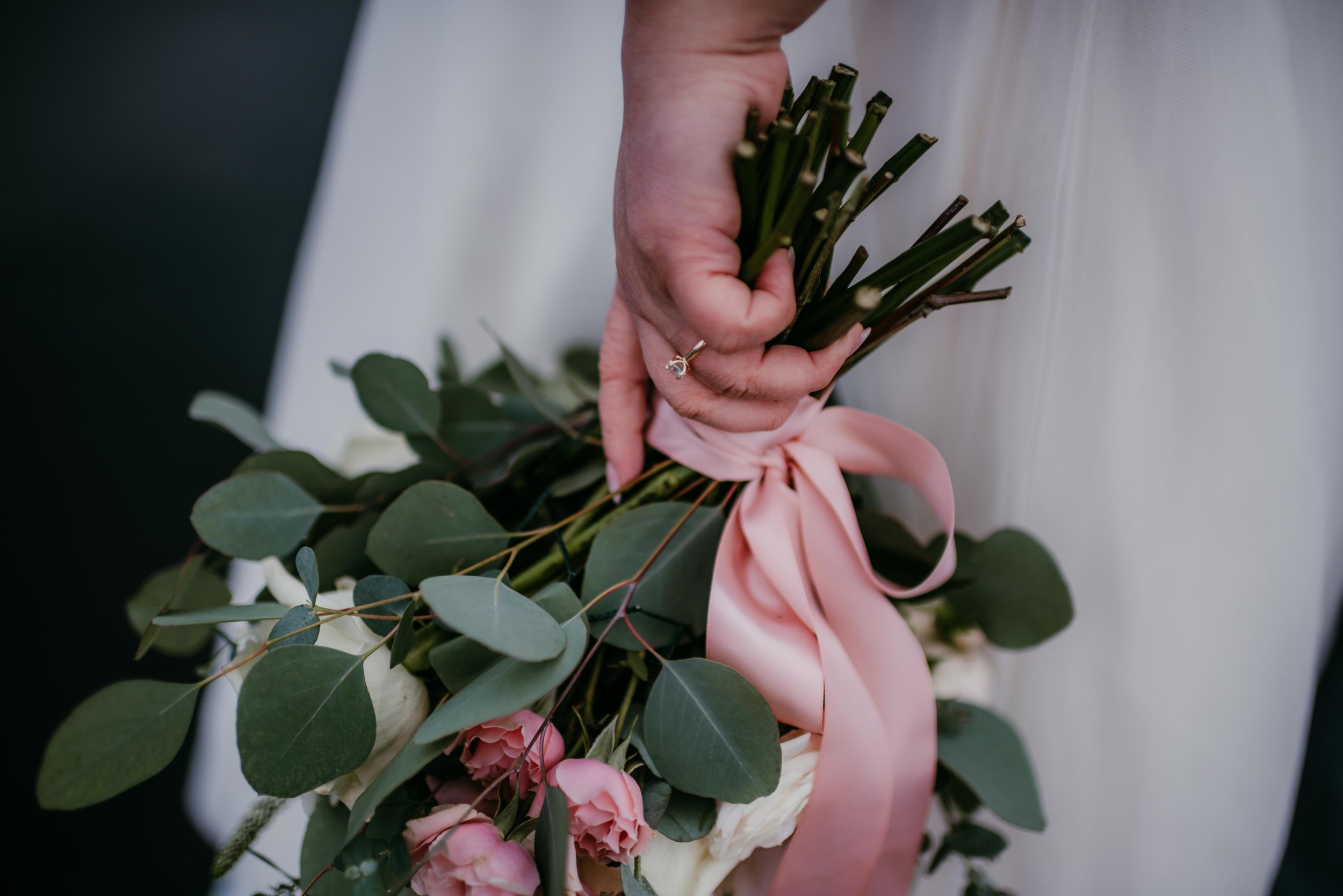 A hand holding a bouquet with green leaves, pink roses, and a pink ribbon.