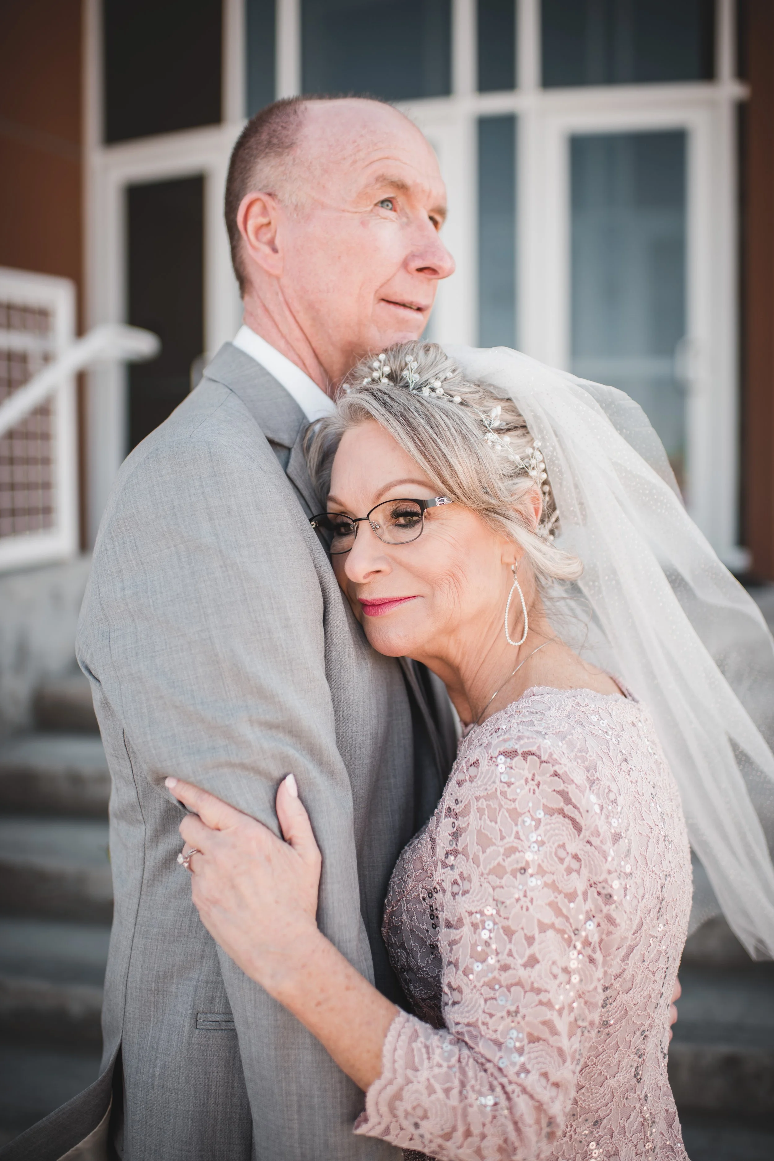 An elderly woman and an elderly man embracing outdoors, the woman wearing a pink lace dress and a veil, and the man in a gray suit, both smiling gently.