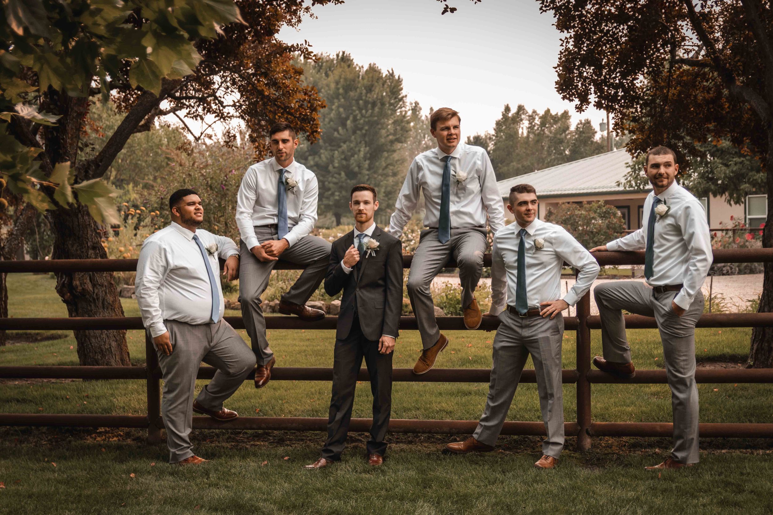 Six men dressed in formal attire posing outdoors near a wooden fence, with trees and a building in the background.