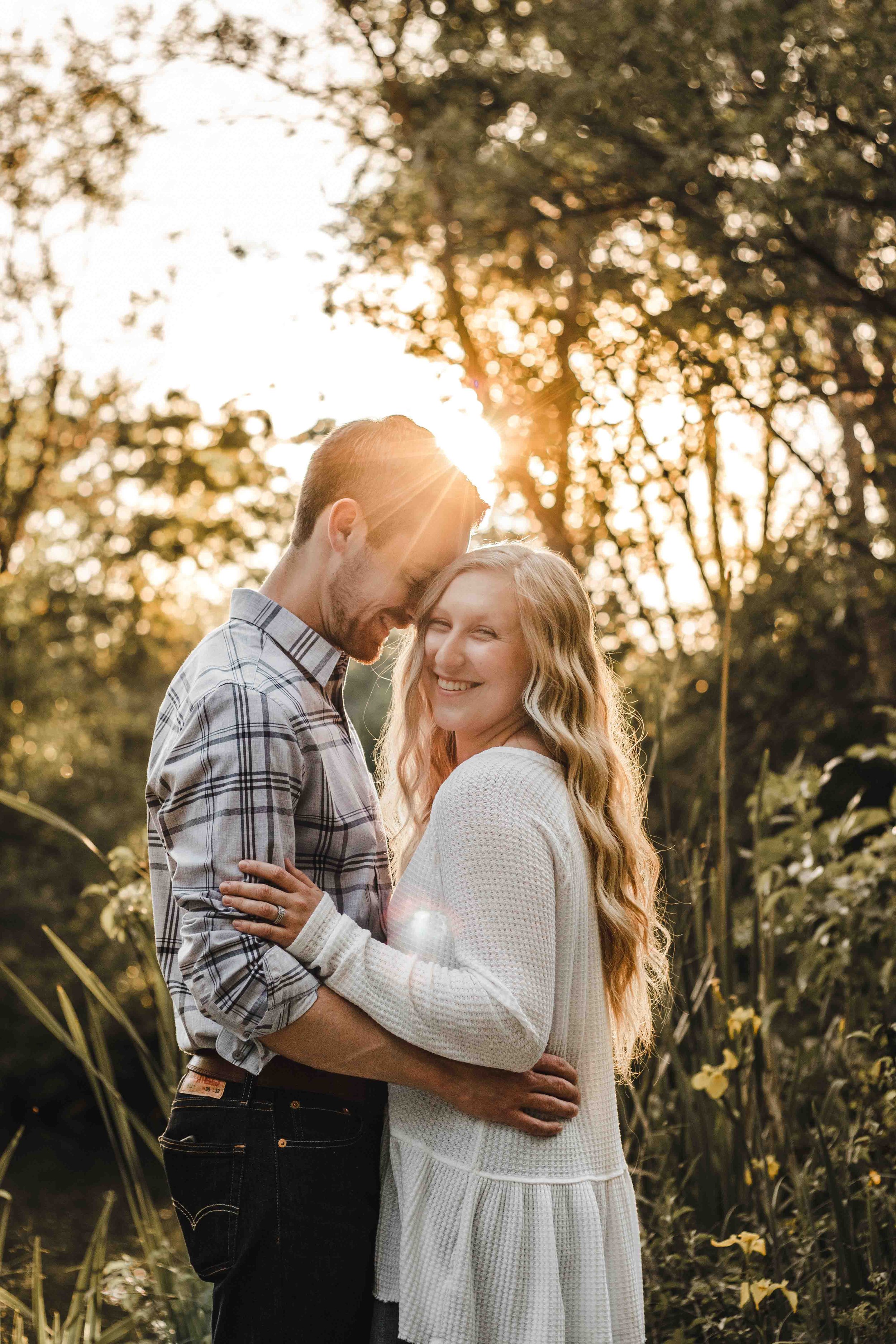 A couple embracing in a sunlit wooded area; the woman smiles while the man leans his forehead against hers, surrounded by trees and plants.