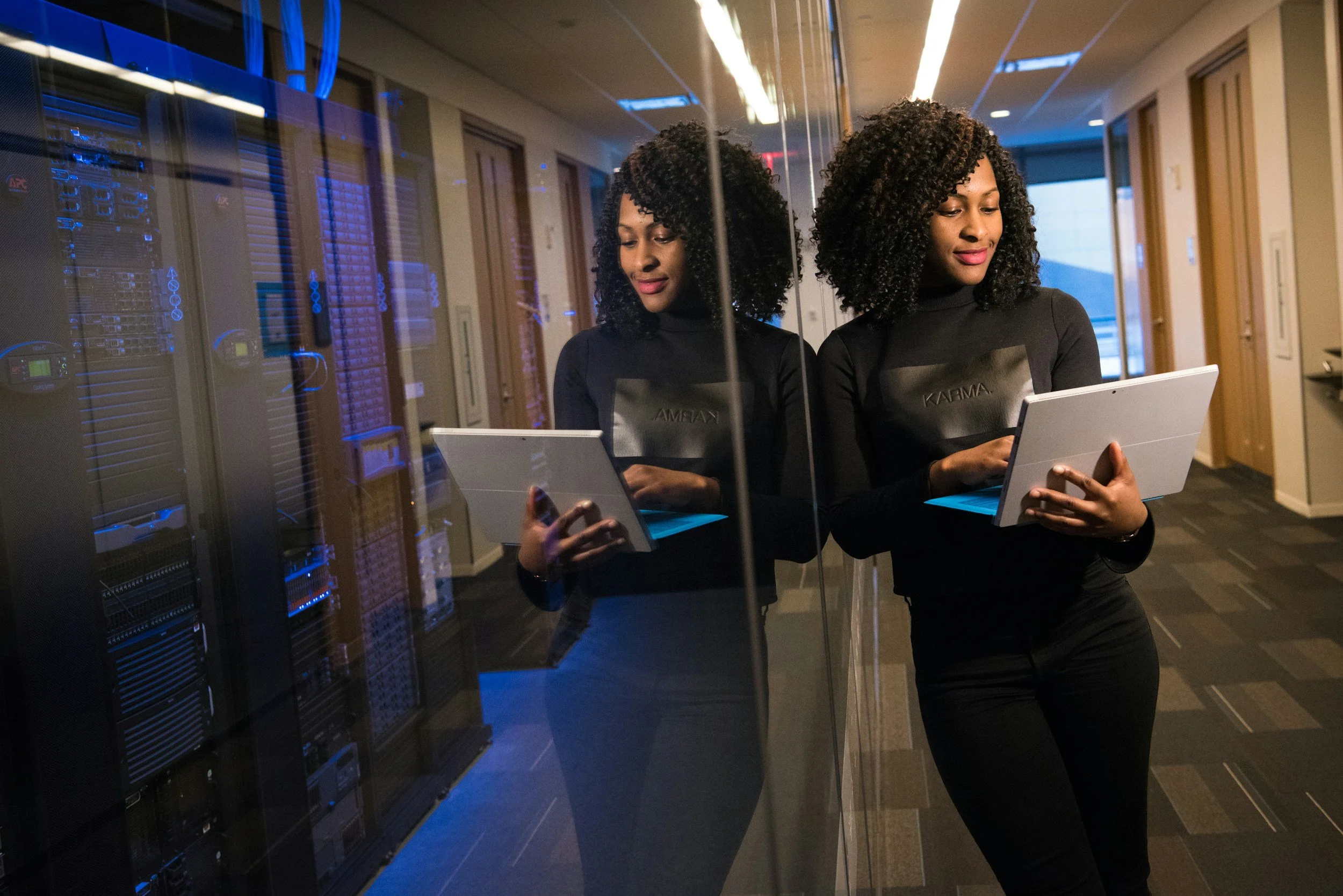 A woman with curly hair in a black long sleeve shirt and black pants standing in a server room with glass walls, holding a tablet, and reflecting in the glass.