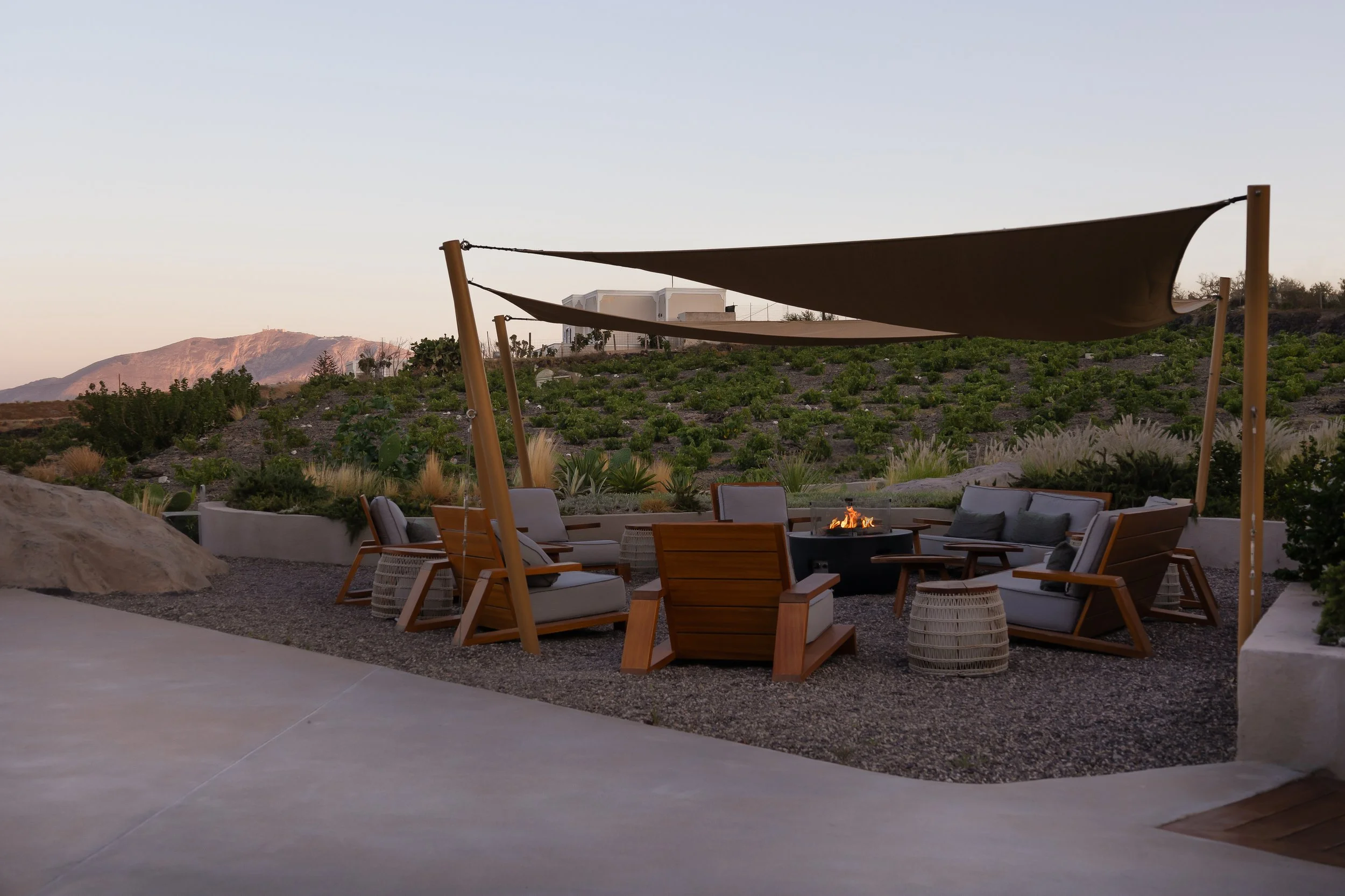 Outdoor seating area with wooden chairs and cushions around a fire pit, shaded by sun sails, with a desert landscape and mountains in the background.
