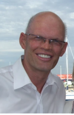 Bald man wearing glasses, smiling outdoors with sky and boats in background