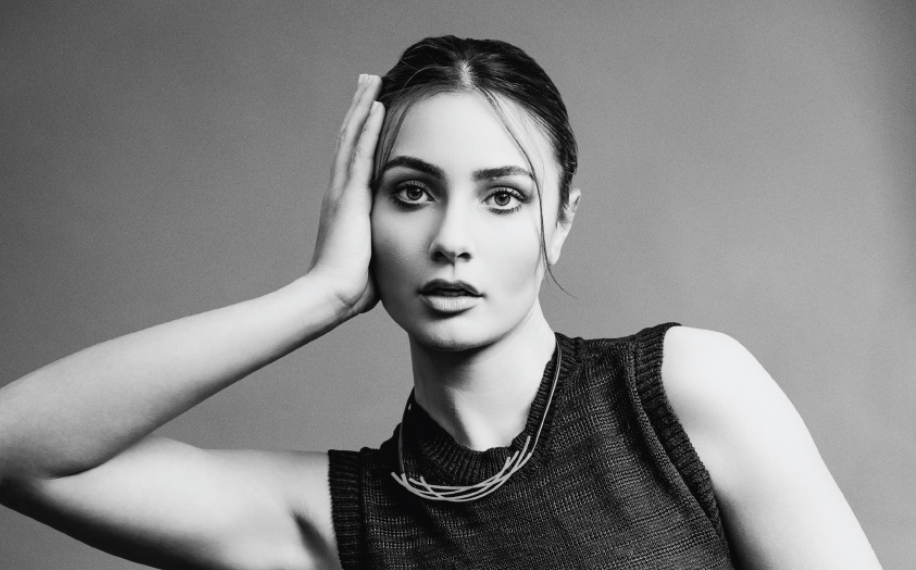 Black and white portrait of a woman posing with one hand on her face, wearing a sleeveless top and a layered necklace.