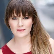 Person with long brown hair and bangs, wearing a red outfit, looking directly at the camera with a blurred urban background.