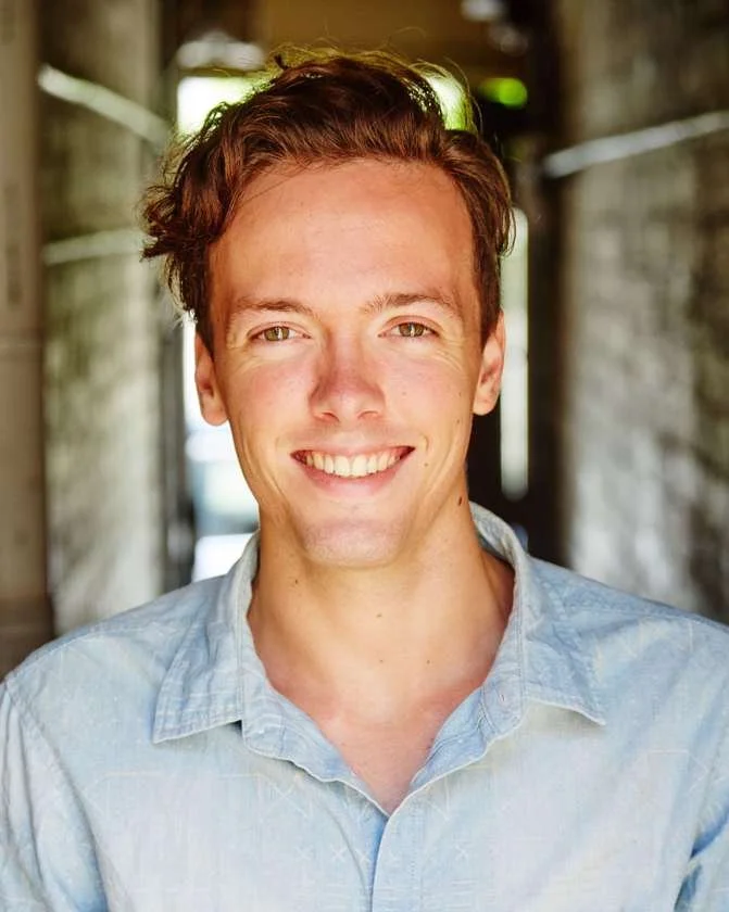 Smiling person in a blue shirt standing in a well-lit hallway.