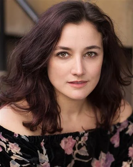 Portrait of a person with dark hair and an off-the-shoulder floral top, looking at the camera.