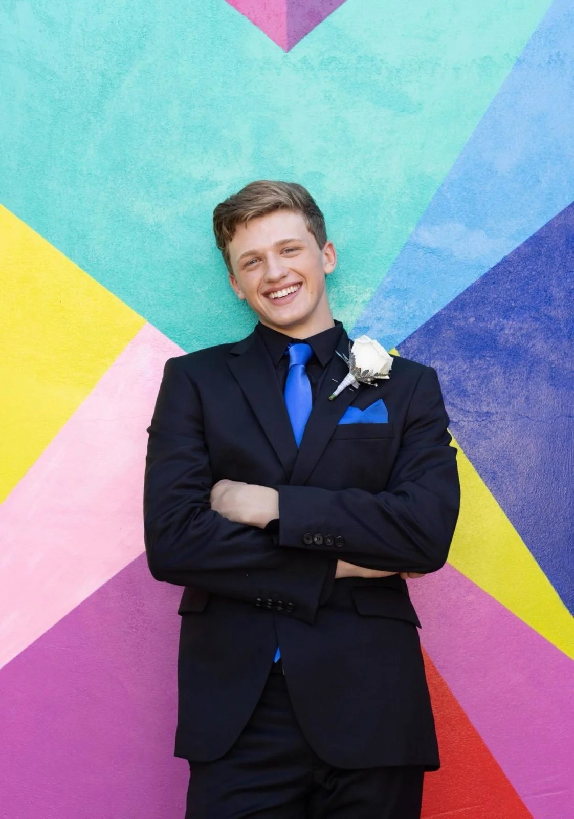 Young man in a black suit with a blue tie and pocket square, smiling against a colorful geometric mural background.