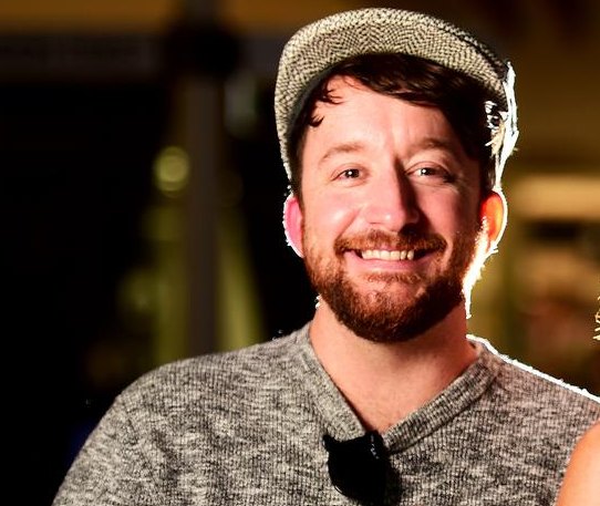 A man wearing a tweed cap and gray sweater, smiling with a blurred background.