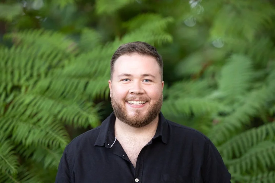 Smiling man in a black shirt standing in front of green foliage.