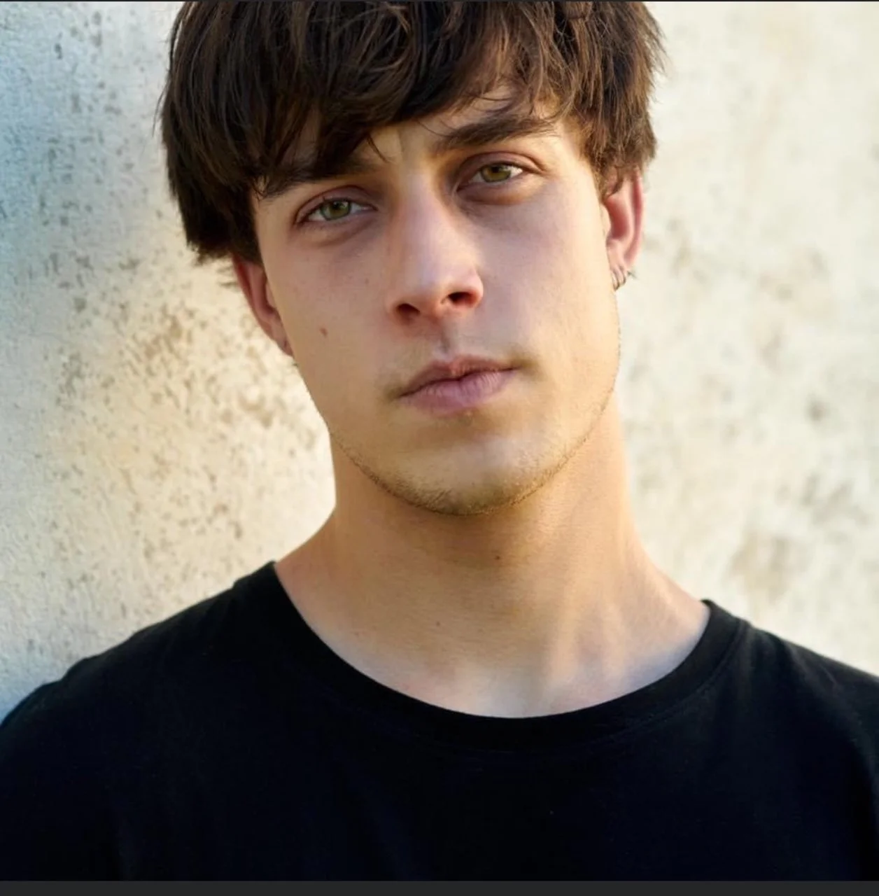Young man with short dark hair, wearing a black shirt, standing against a textured wall, looking directly at the camera.