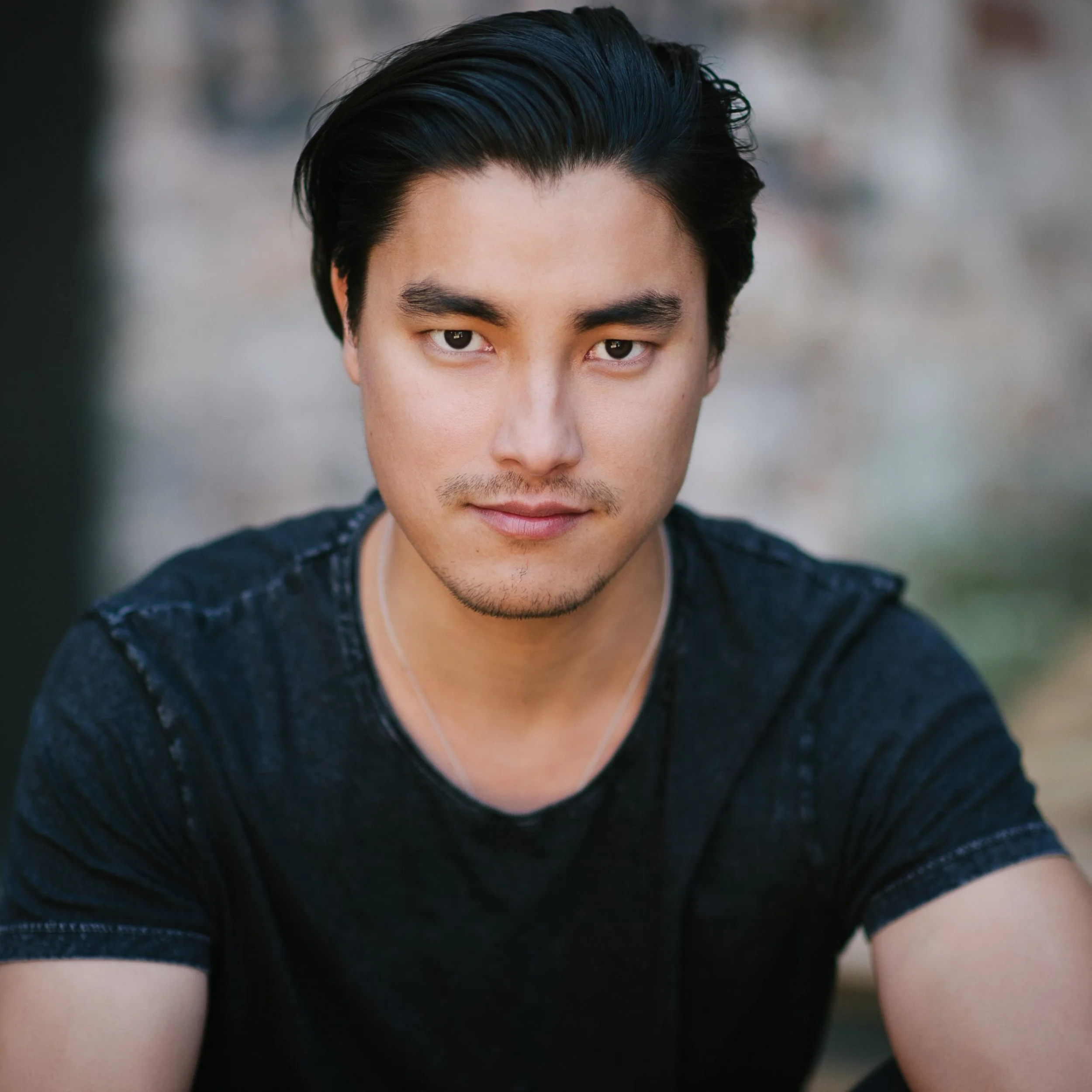 Person with short black hair wearing a dark t-shirt, looking at the camera, with a blurred background.