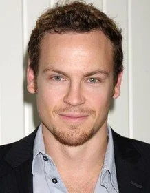 Portrait of a man with short curly hair and a light beard wearing a suit jacket and shirt.