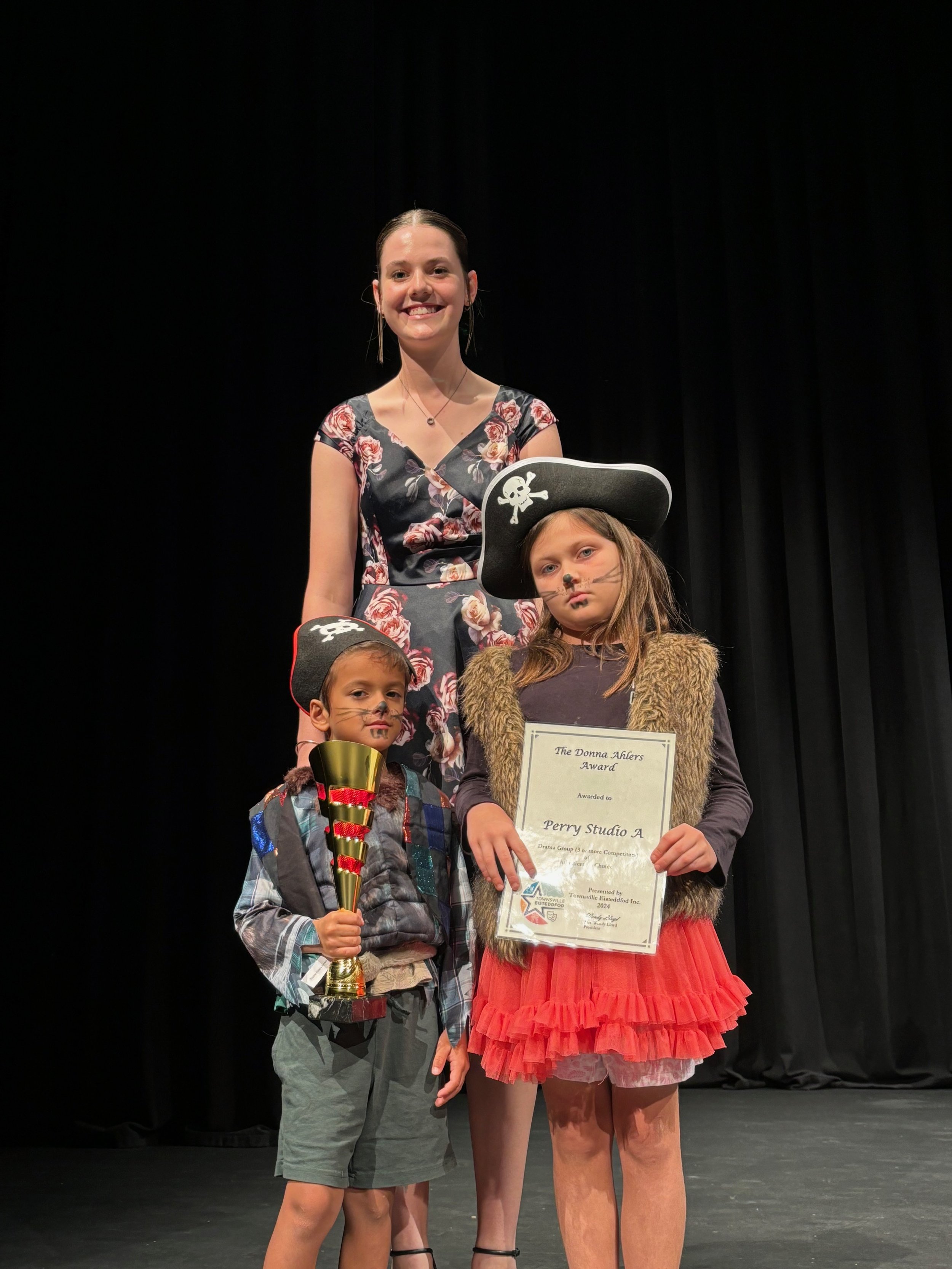 Two children in pirate costumes and painted faces stand in front of a smiling woman on stage. The boy holds a trophy, and the girl holds a certificate with 'The Donna Adlers Award' printed on it. The backdrop is a dark curtain.